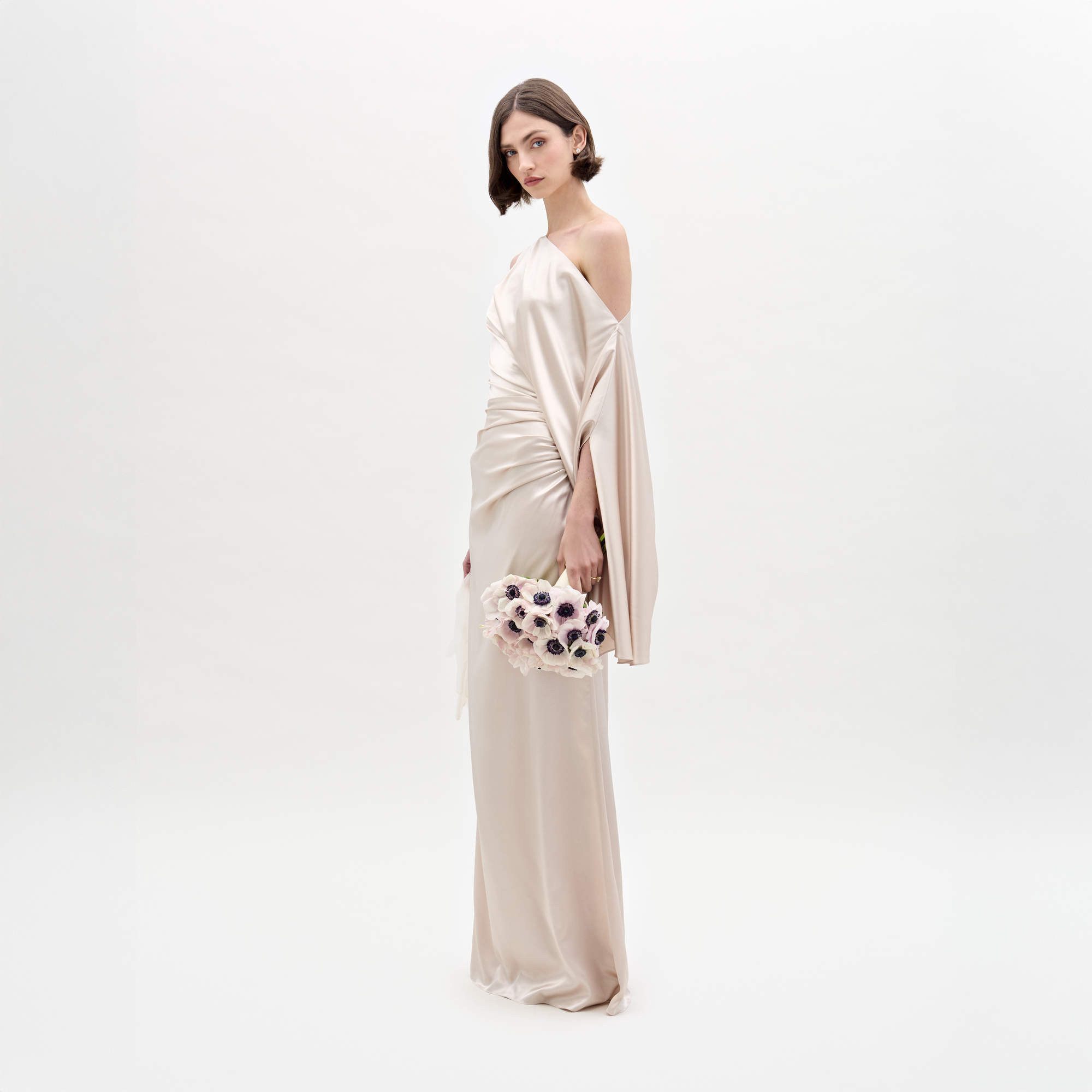 Una mujer con el Vestido Dotty, un vestido largo maxi de tela italiana con escote asimétrico, está de pie mirando hacia adelante sosteniendo un ramo de flores pálidas con centros oscuros sobre un fondo blanco liso.