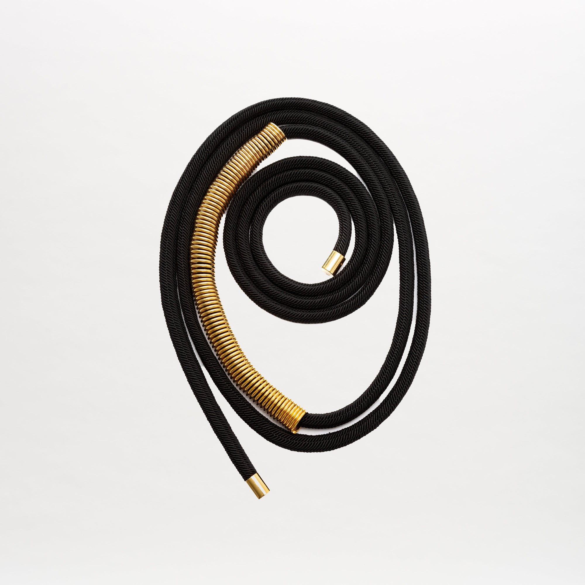 El cinturón Dunia, un cinturón de cordón negro con espirales de metal dorado, se enrolla en una espiral suelta sobre un fondo blanco liso.