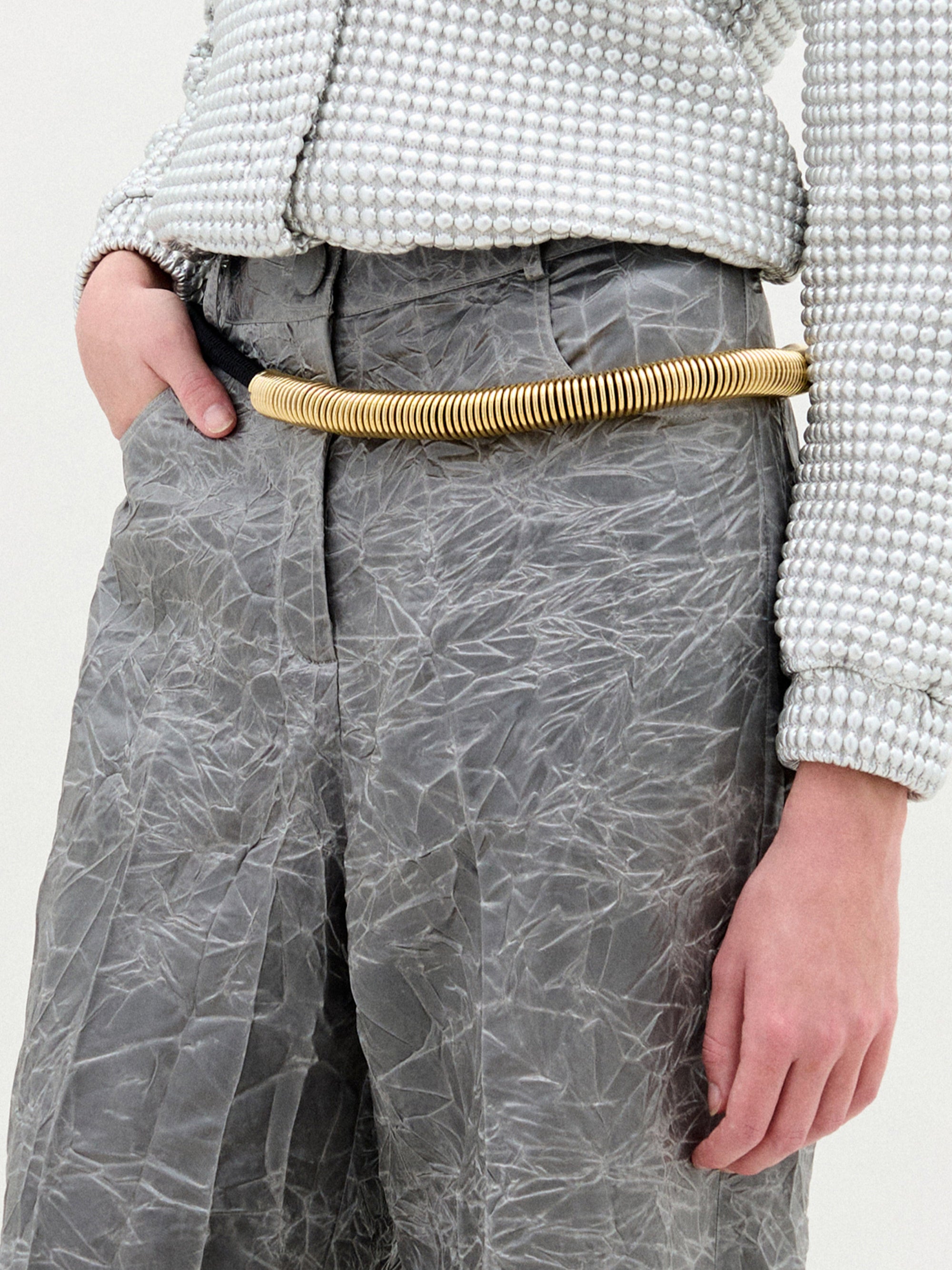 Con unos pantalones grises arrugados y una camisa blanca texturizada con pequeños lunares en relieve, esta persona lleva como accesorio el cinturón Dunia, un grueso cinturón dorado hecho a mano en Colombia, que destaca mientras su mano izquierda descansa en un bolsillo.