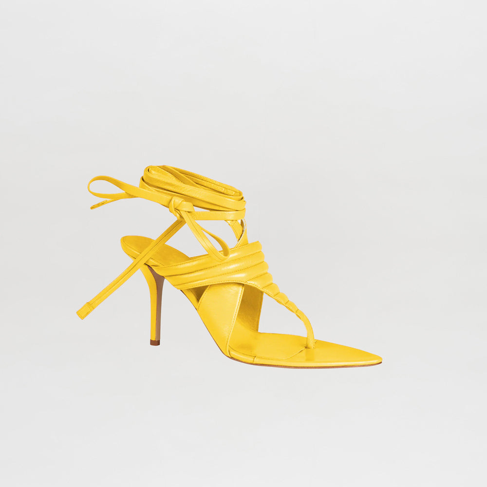 Tacones Dalila Limoncello es una sandalia de tacón alto de color amarillo que presenta una punta puntiaguda, un tacón de aguja fino, un diseño de tiras en la parte superior y cordones largos que envuelven el tobillo, todo ello sobre un fondo blanco liso.