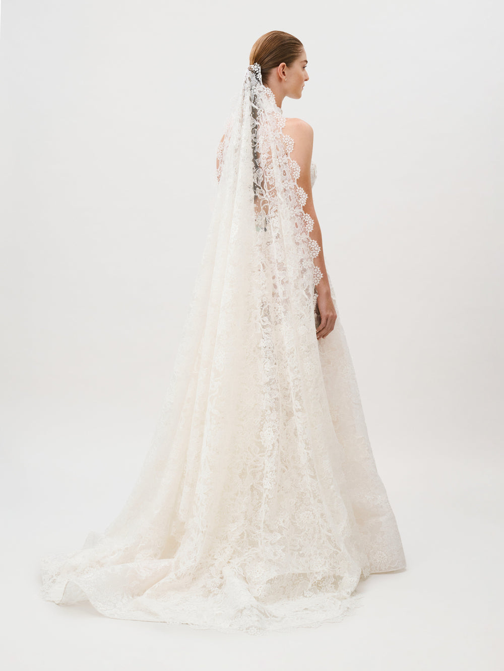 A bride in the Danita Dress—a white lace wedding gown crafted from luxurious Italian fabric—stands with her back to the camera, a long, elegant lace veil trailing behind her against a plain white background: a true vision of timeless beauty from Colombia Atelier.