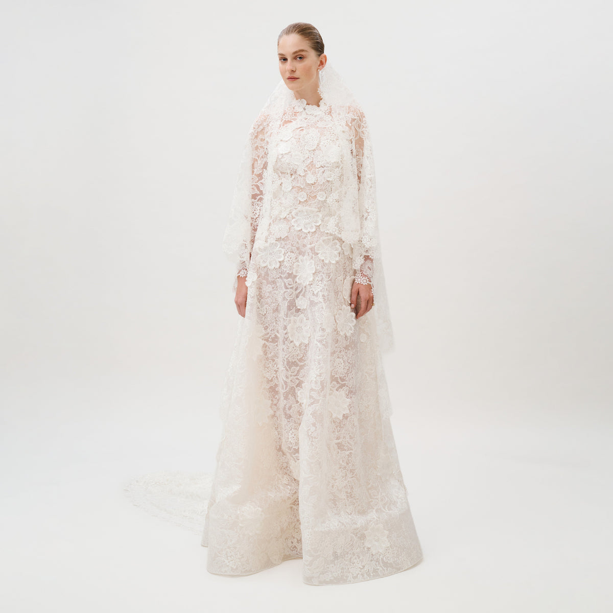 Dressed in the Danita Dress—a long, white, lace wedding gown adorned with floral patterns and crafted from exquisite Italian fabric by Colombia Atelier—a woman stands against a plain white background. Her timeless bridal look is completed with a matching lace veil.