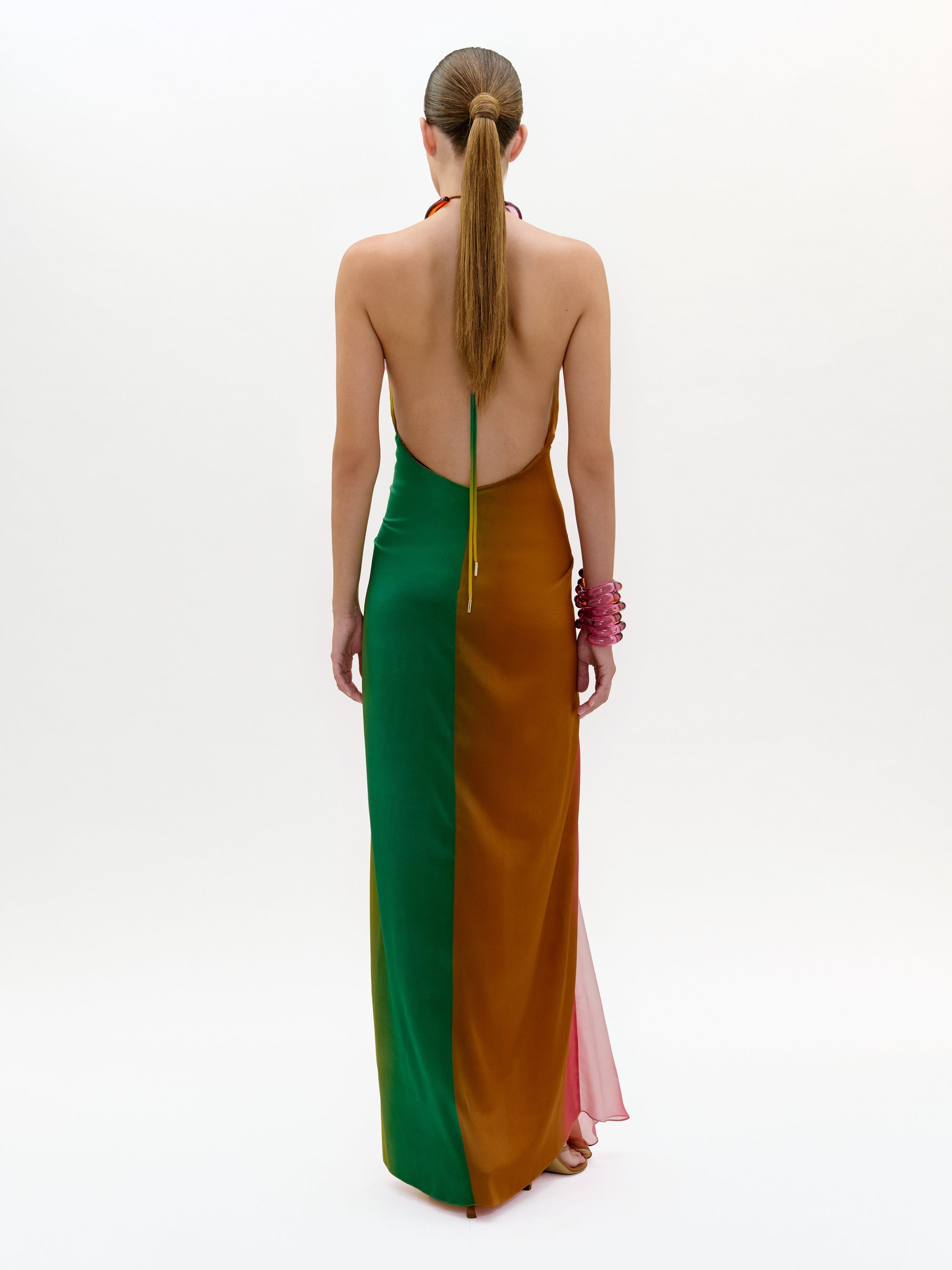 Una persona con el pelo largo recogido en una coleta aparece de espaldas con el vestido Dixon, un maxi largo con escote halter y espalda descubierta en tejido italiano verde y naranja con un panel lateral rosa transparente, y una pulsera rosa.