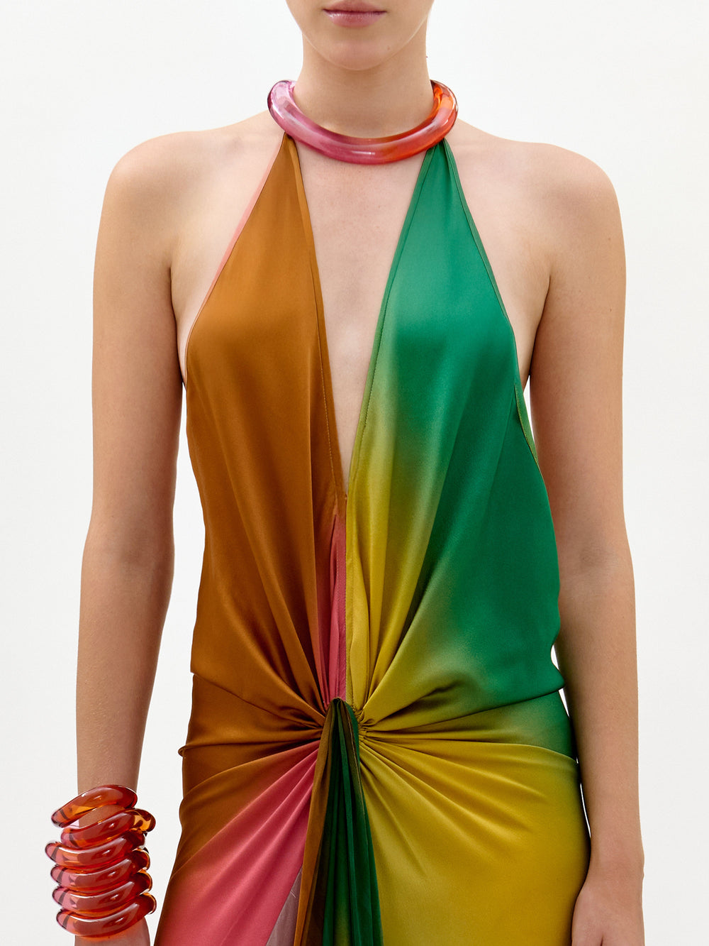 Una persona lleva el vestido Dixon, una maxi columna sin mangas y con un profundo escote en V, confeccionado en tejido italiano en un degradado de naranja, verde, amarillo y rosa, fruncido en la cintura y peinado con una gargantilla translúcida gruesa y pulseras a juego.
