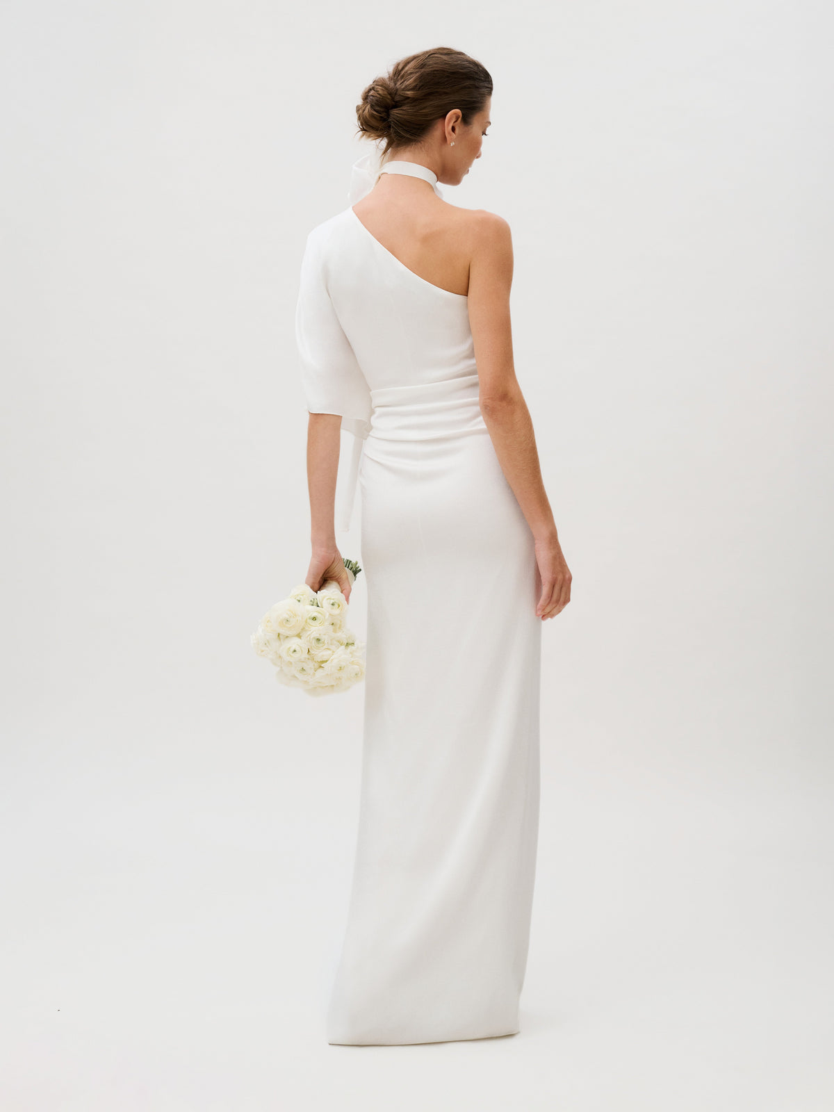 A woman stands with her back to the camera against a plain light background, holding a bouquet of white flowers and wearing the Doli Dress, a one-shoulder white hammered satin gown.