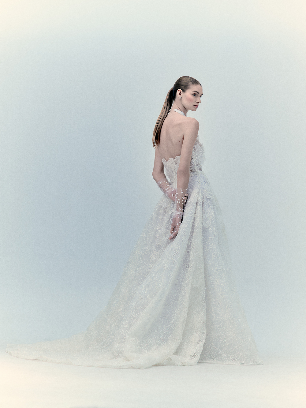Una mujer de perfil luce el Vestido Dorothy, un vestido de encaje blanco sin tirantes con apliques florales, una larga cola y guantes transparentes. Lleva el cabello liso y recogido, sobre un fondo con un suave degradado.