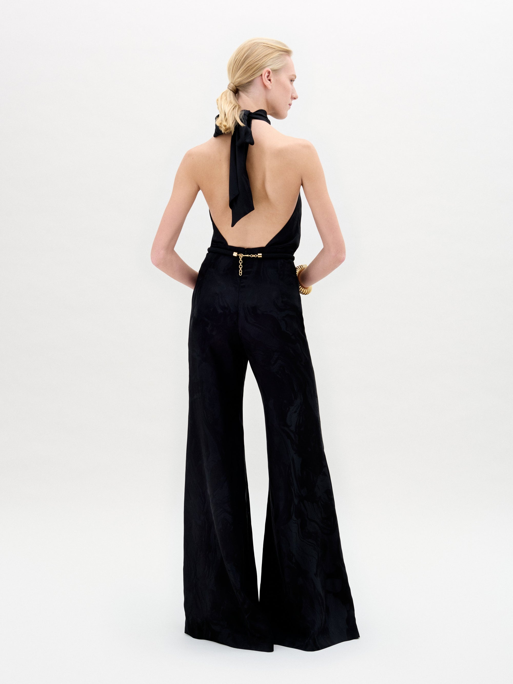 Una mujer con el pelo rubio recogido en una coleta baja, de espaldas a la cámara, lleva el mono Enara -con top negro de escote halter y pantalones de cintura alta y pernera ancha de tela italiana- sobre un fondo claro y liso.