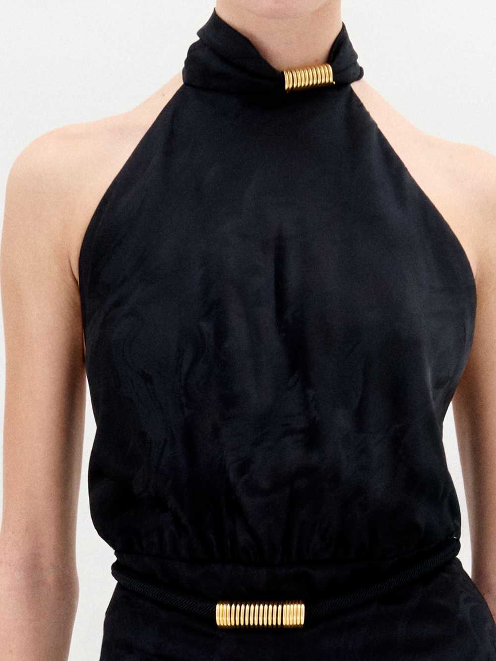 Una persona lleva puesto el mono Enara, una prenda sin mangas de escote halter con tejido fruncido en el cuello y la cintura, ambos realzados por detalles cilíndricos dorados. Hecho en Colombia, sobre un fondo claro y liso.