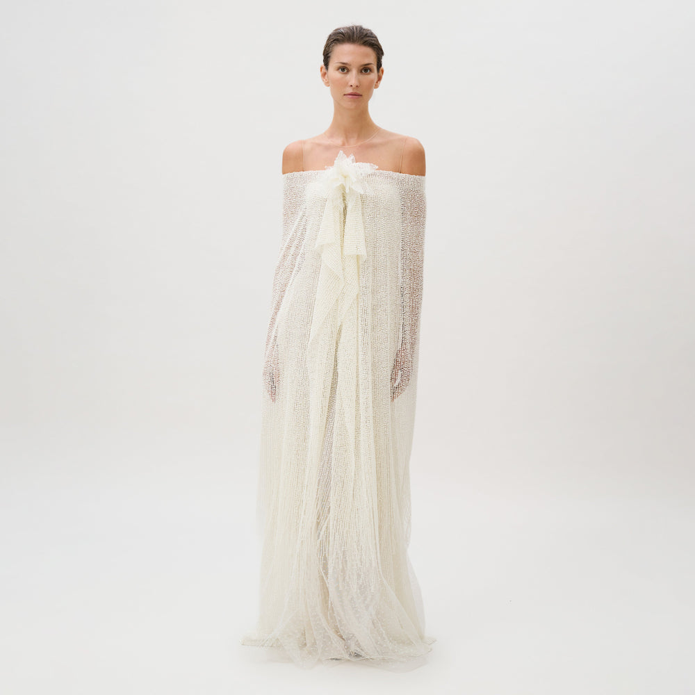 A woman stands against a plain white background, wearing the Edna Dress—an off-the-shoulder, textured white gown crafted from luxurious fabric with long, flowing layers and delicate sheer accents. This exquisite piece is masterfully made in Italy.