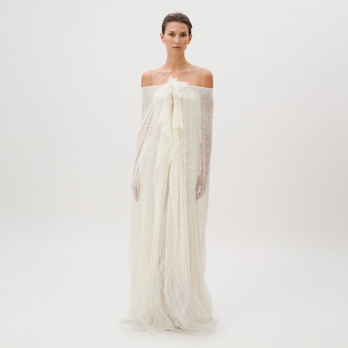 A woman stands against a plain white background, wearing the Edna Dress—an off-the-shoulder, textured white gown crafted from luxurious fabric with long, flowing layers and delicate sheer accents. This exquisite piece is masterfully made in Italy.