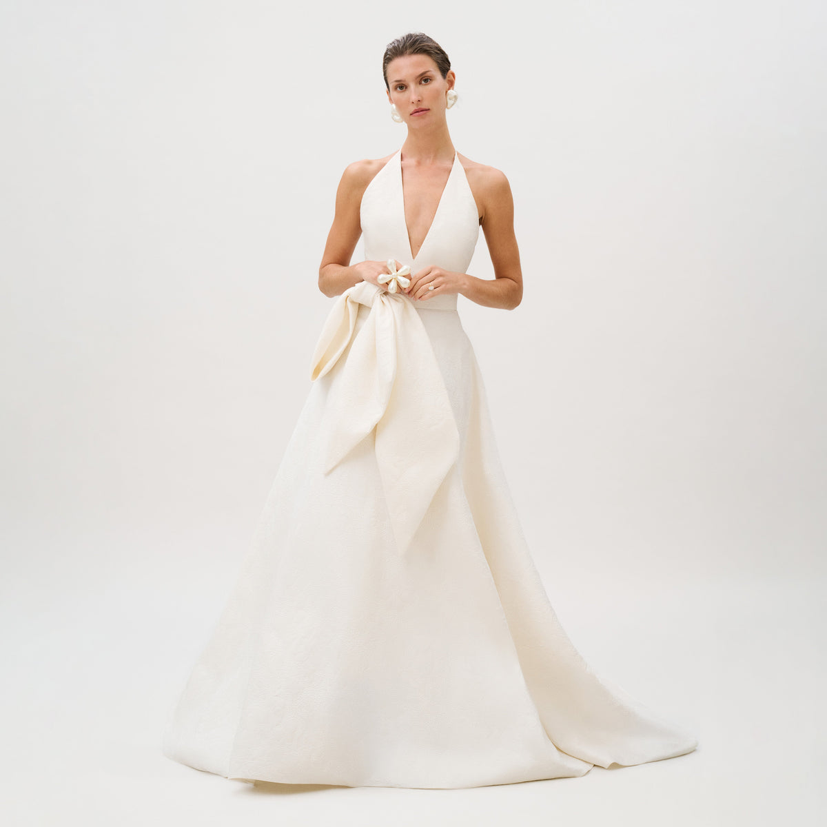 Dressed in the exquisite Elsa Dress—a sleeveless, floor-length white gown from our Atelier collection, crafted from luxurious Italian fabric and featuring a deep V neckline with a large bow at the waist—the woman stands against a plain light background.