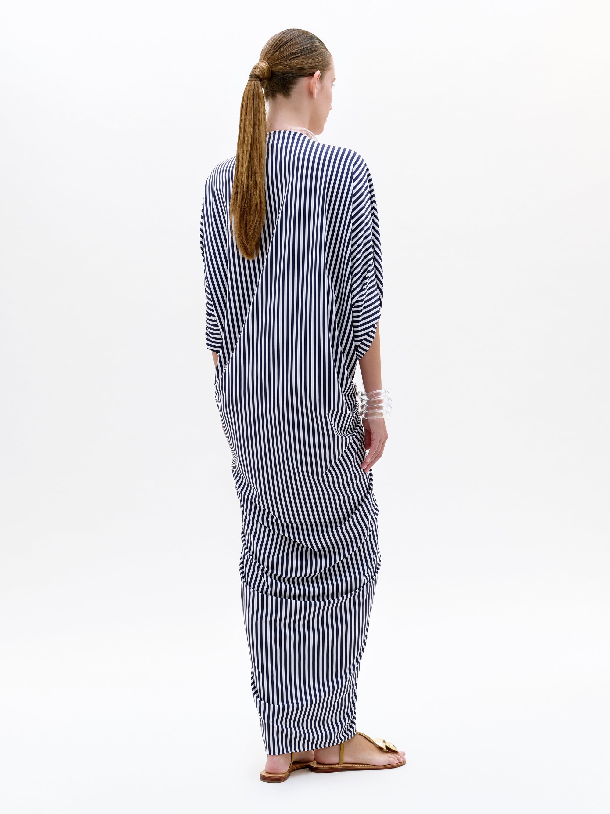 Una persona con el pelo largo recogido en una coleta se coloca de espaldas, con la túnica Fiore -rayas verticales blancas y negras a modo de maxivestido- y sandalias planas, sobre un fondo blanco liso.