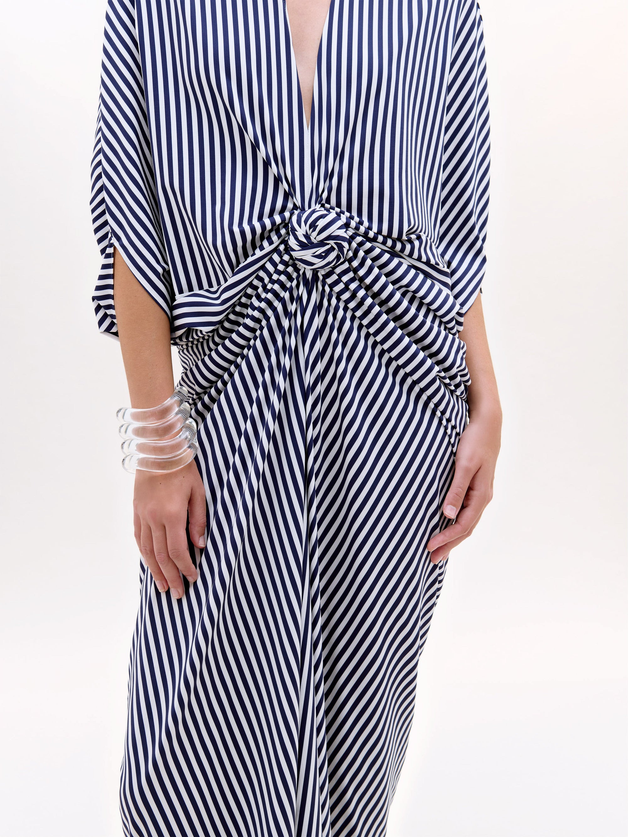 Se ve a una persona con la Túnica Fiore, un maxivestido de rayas verticales azules y blancas con cintura de nudo retorcido, escote en V y un brazalete de trozos transparentes. No se ve la cabeza; el fondo es liso y claro.
