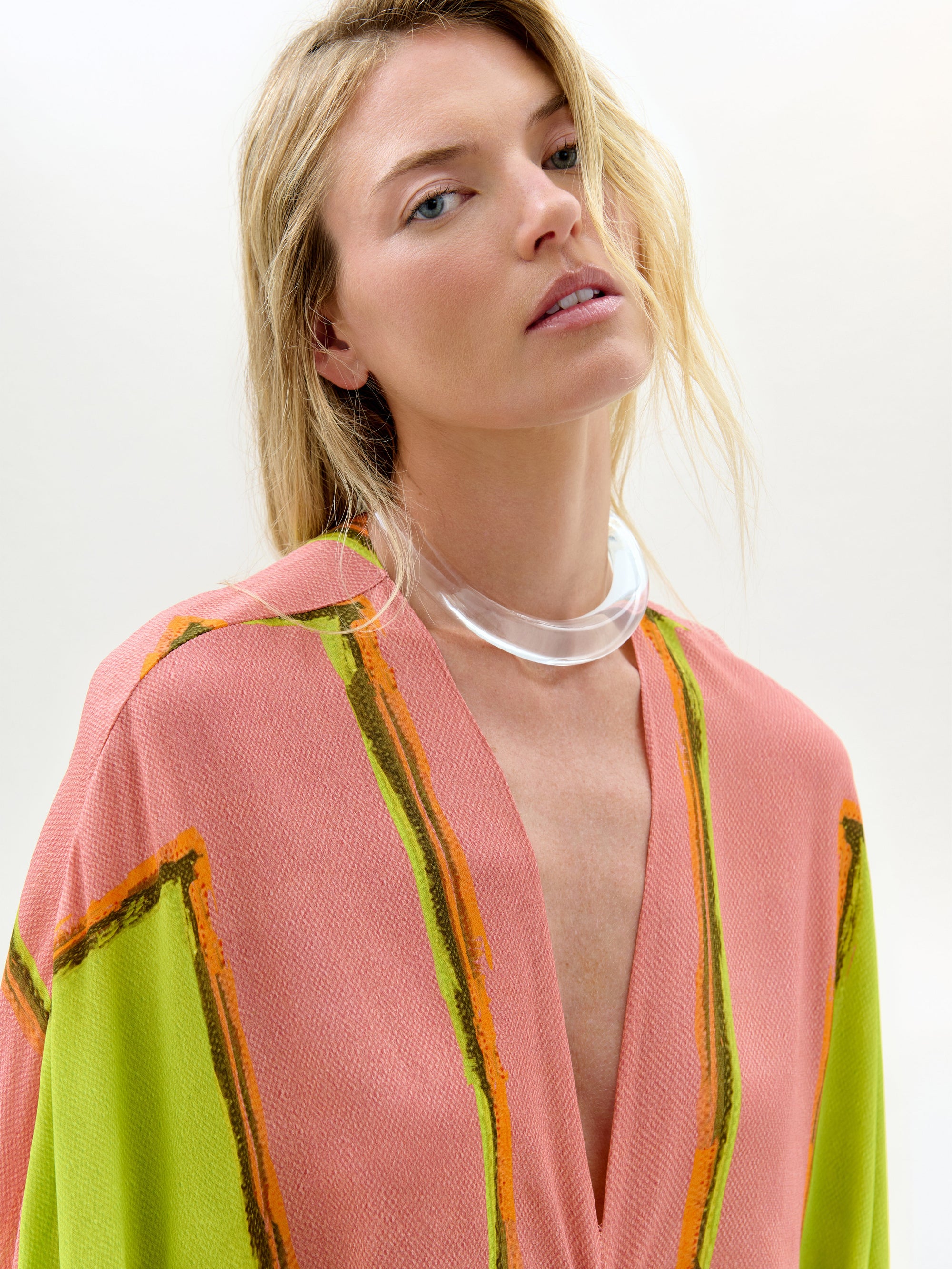 Una mujer de pelo largo y rubio lleva la Túnica Fiore, una maxi holgada en rosa y verde con toques naranjas, confeccionada en tejido italiano. La combina con un collar grueso transparente y mira hacia arriba sobre un fondo liso.