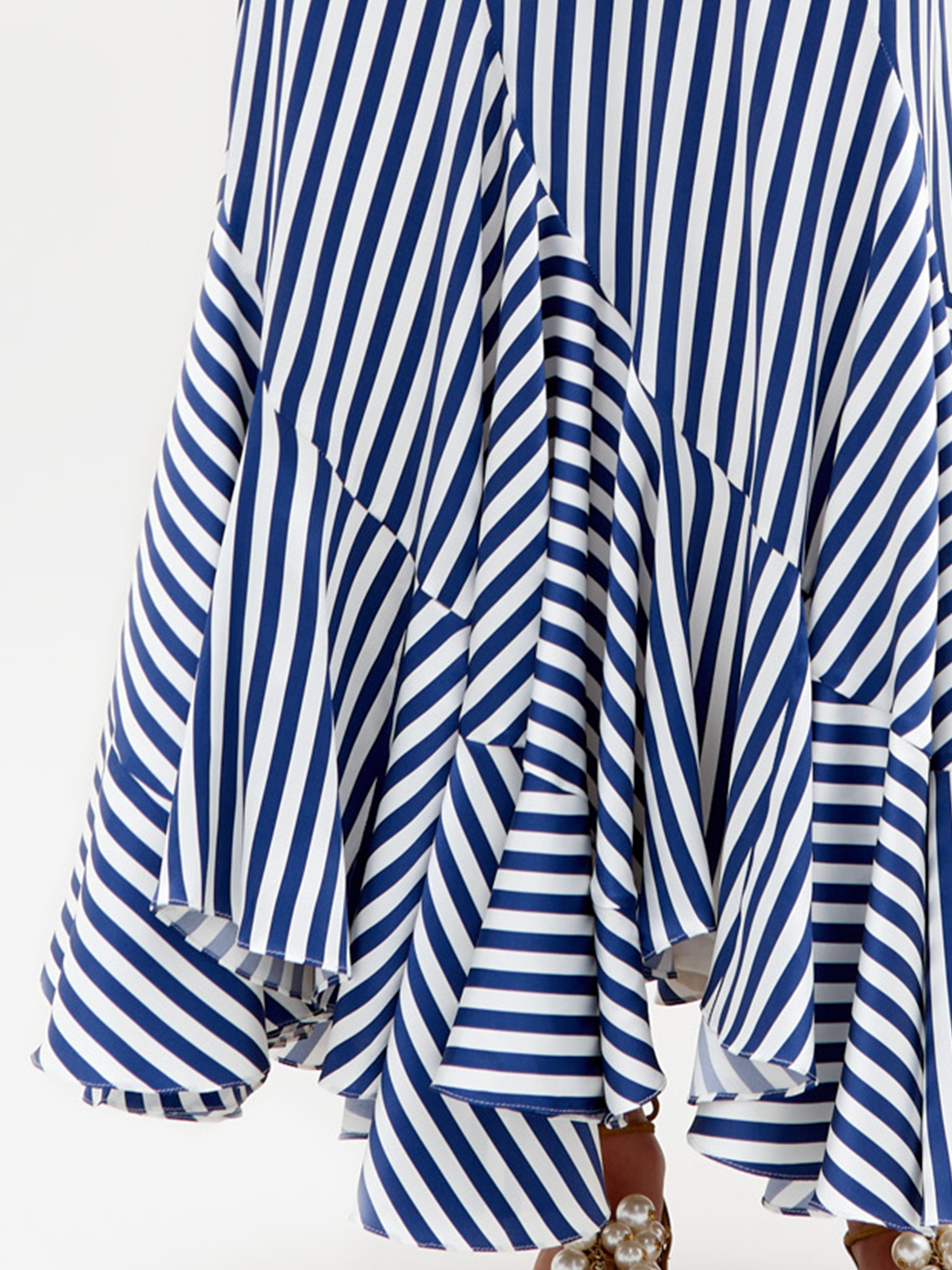 Primer plano de Falda Florina, una falda con volantes de rayas azules y blancas confeccionada en textil italiano con rayas diagonales. Presenta un dobladillo asimétrico con una sutil silueta sirena y bordes de volantes ligeramente rizados; las perlas en la parte inferior sugieren que pueden ser parte del calzado.
