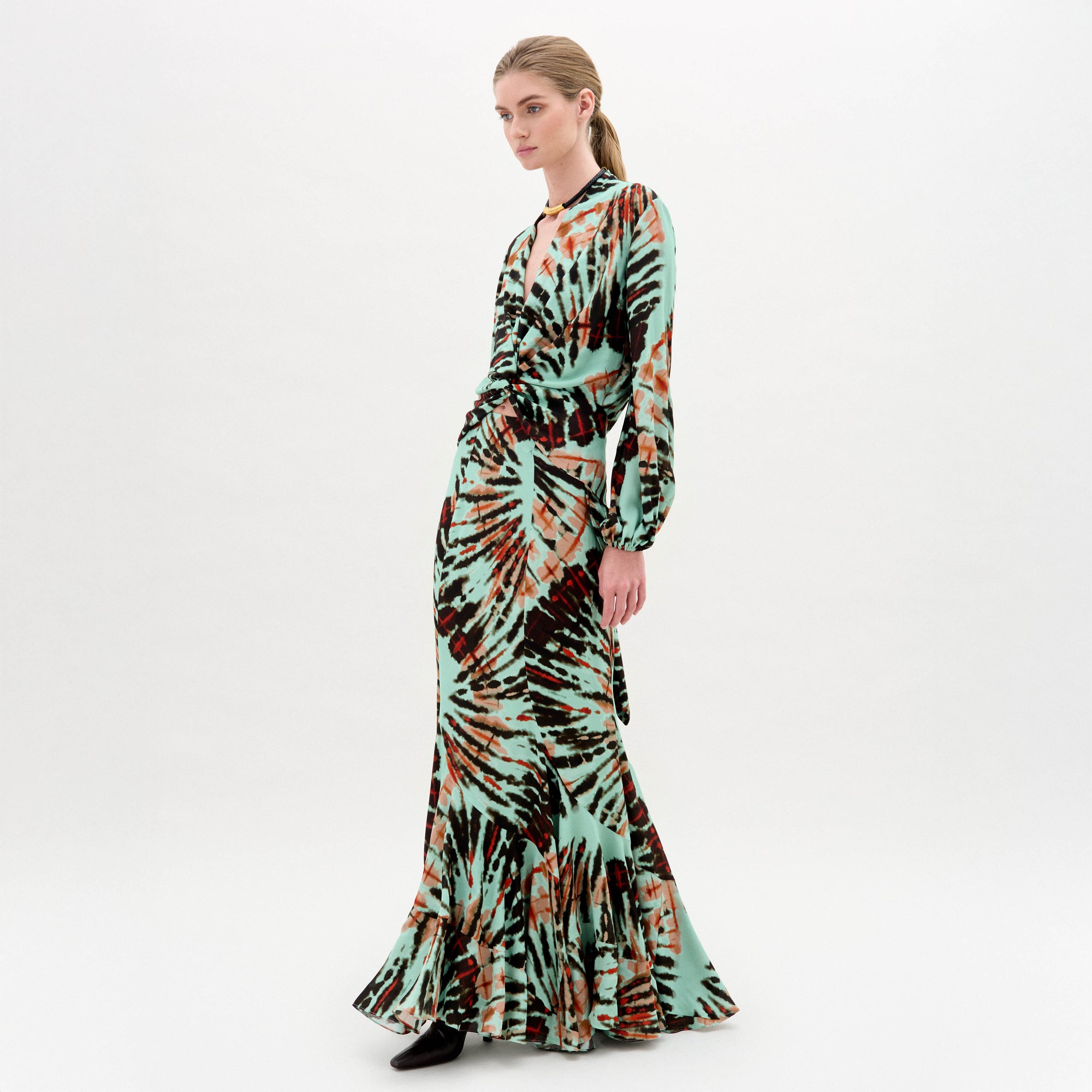 Una mujer con el pelo rubio recogido lleva la falda Florina, una maxi larga hasta el suelo con estampados tie-dye en verde, marrón y naranja, combinada con zapatos negros sobre fondo blanco. Hecho en Colombia.
