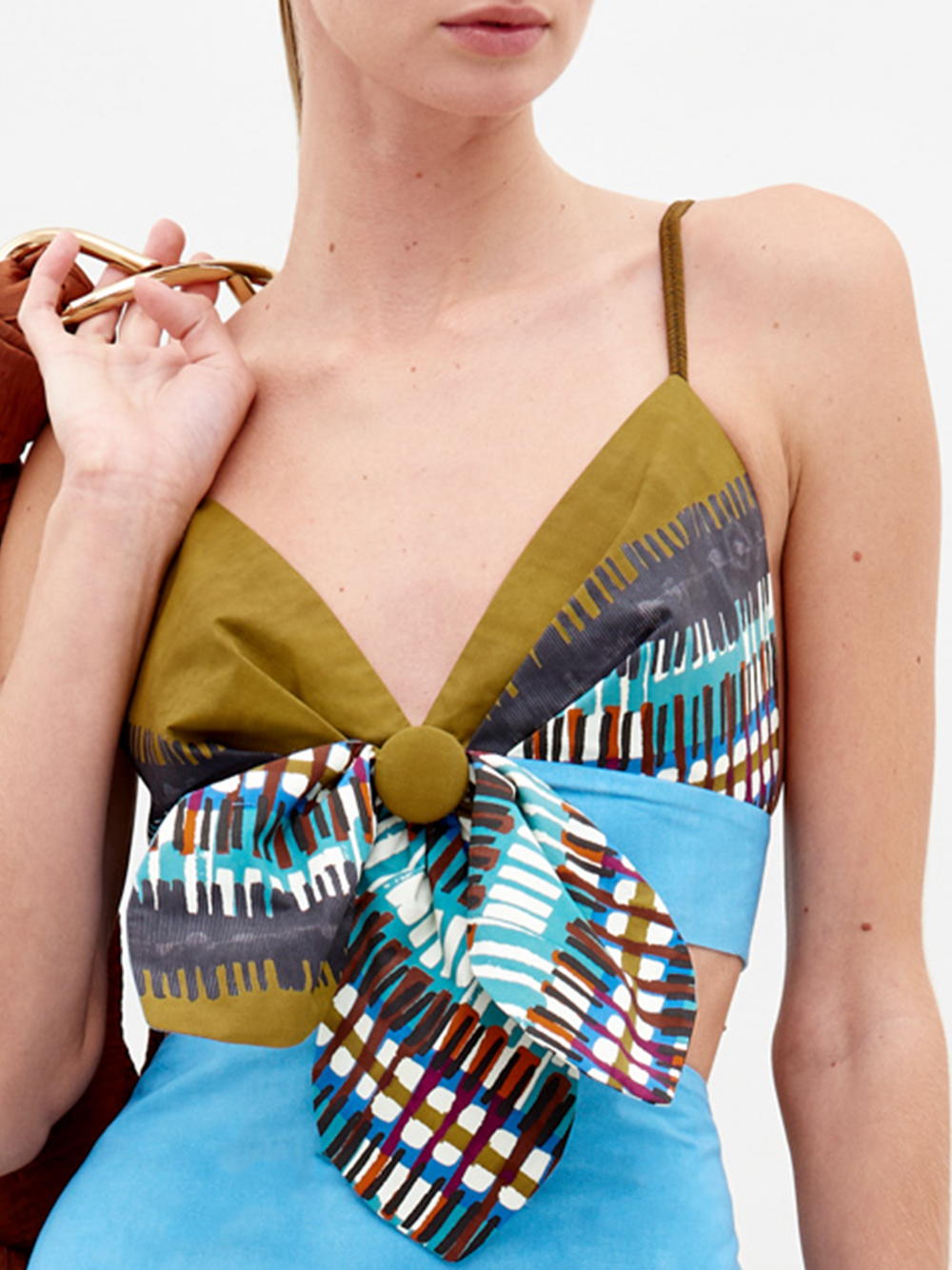 Una mujer lleva el Top Flor, un vestido corto sin mangas con tirantes finos, escote profundo en pico, motivos geométricos verde oliva y multicolor, un gran lazo en el busto y tejido italiano azul claro bajo el pecho.