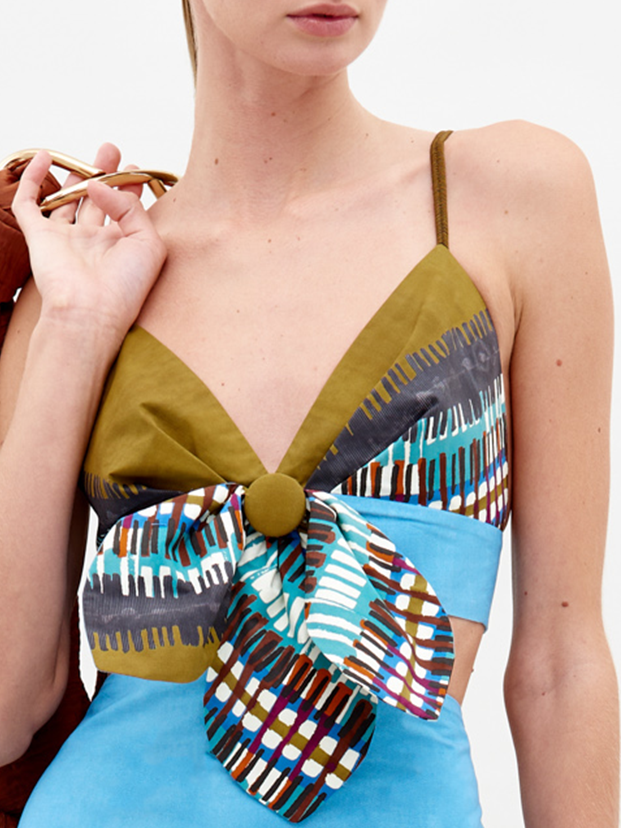 Una mujer lleva el Top Flor, un vestido corto sin mangas con tirantes finos, escote profundo en pico, motivos geométricos verde oliva y multicolor, un gran lazo en el busto y tejido italiano azul claro bajo el pecho.