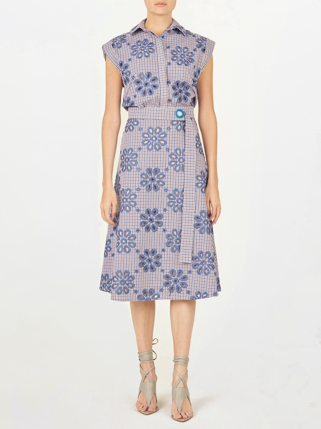 Una persona lleva el Blusa Fosca Navy Floral, con estampado floral azul sobre cuadros rojos y blancos, ceñido a la cintura y combinado con sandalias de cordones gris claro.