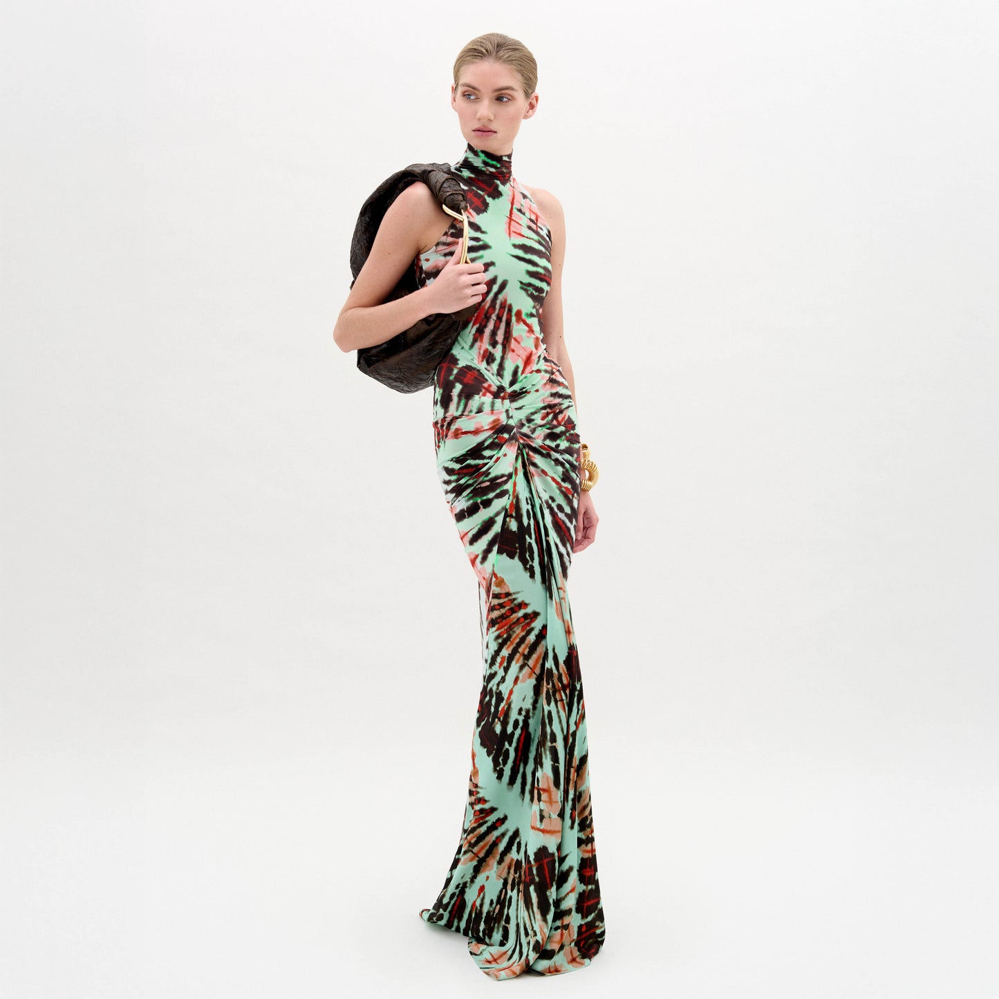 Una mujer posa sobre un fondo blanco con el vestido Frances, un maxi sin mangas hasta el suelo con un llamativo estampado multicolor. Lleva una prenda de textura oscura al hombro y una pulsera gruesa. Hecho en Colombia.