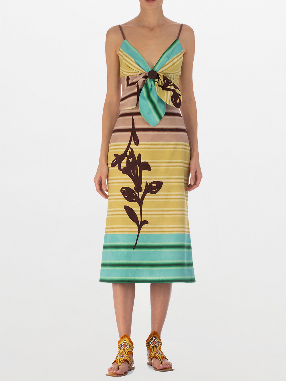 Una mujer sobre fondo blanco lleva el Falda Atira Meadow View, un vestido floral multicolor sin mangas en tonos amarillos, marrones y verdes. Lo combina con sandalias de tiras con cuentas y lleva el pelo recogido.