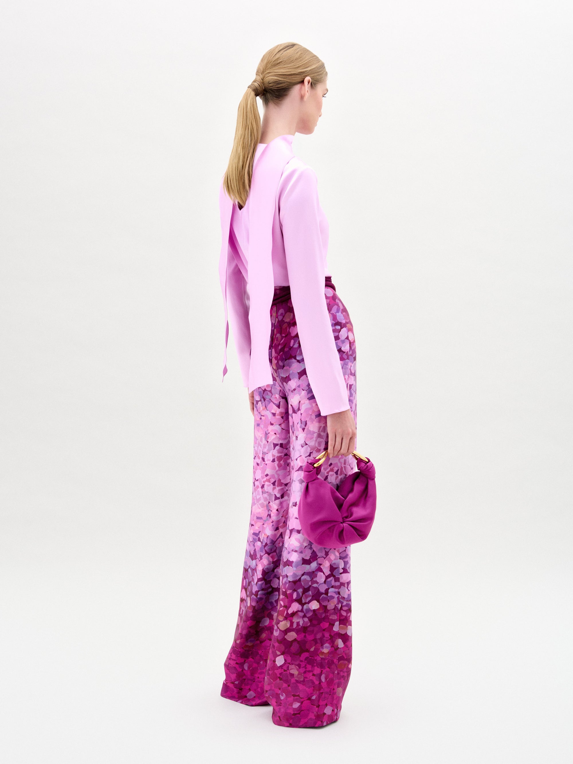 Una mujer con el pelo rubio recogido en una coleta mira hacia otro lado, lleva un top rosa claro y un pantalón Como de talle alto con un estampado rosa y morado en tela italiana, y sostiene un bolso rosa sobre un fondo blanco liso.