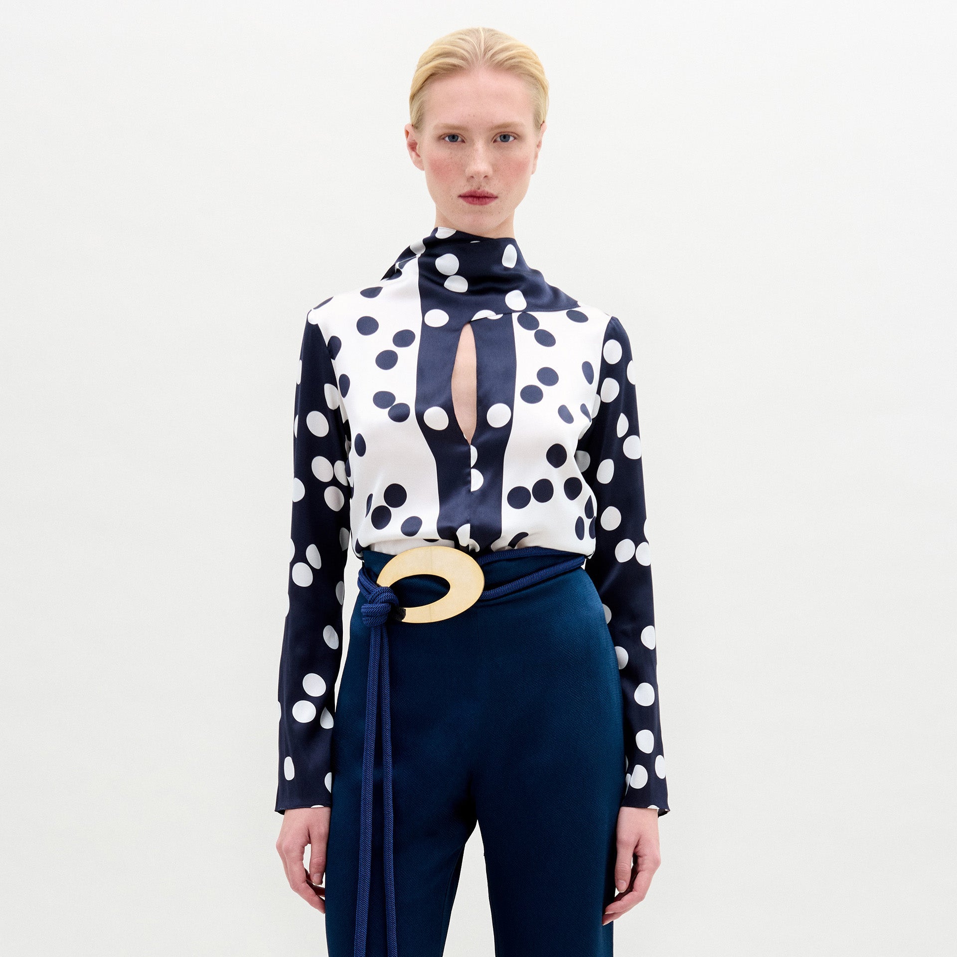 Una persona, sobre un fondo blanco liso, lleva la blusa Gaby, un top de seda de manga larga con lunares marinos y blancos, combinado con un pantalón azul oscuro de talle alto y un cinturón ancho con una gran hebilla circular.