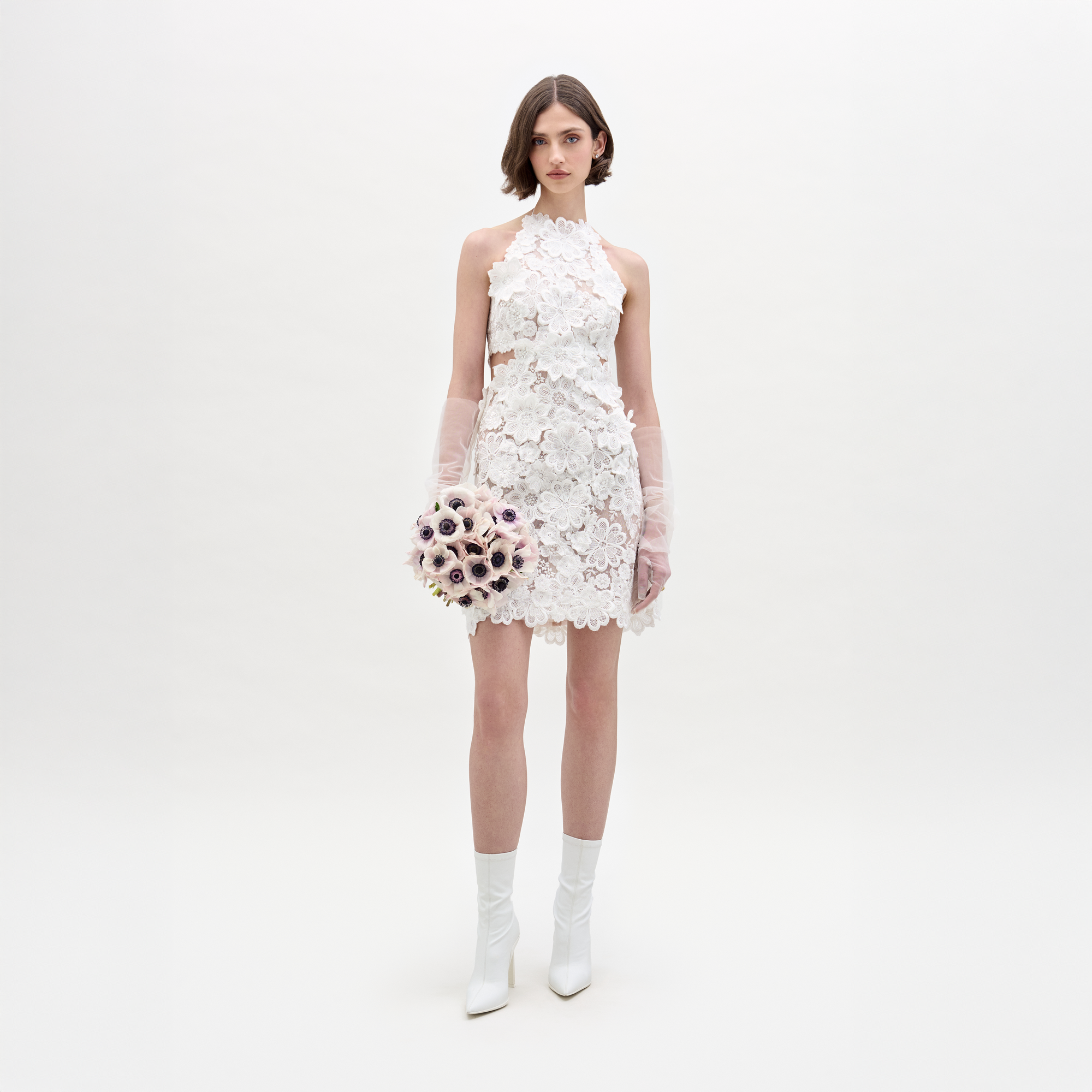 Una mujer aparece sobre un fondo blanco liso, vistiendo el Vestido Garazi, guantes transparentes, botines blancos y sosteniendo un ramo de flores de colores claros.