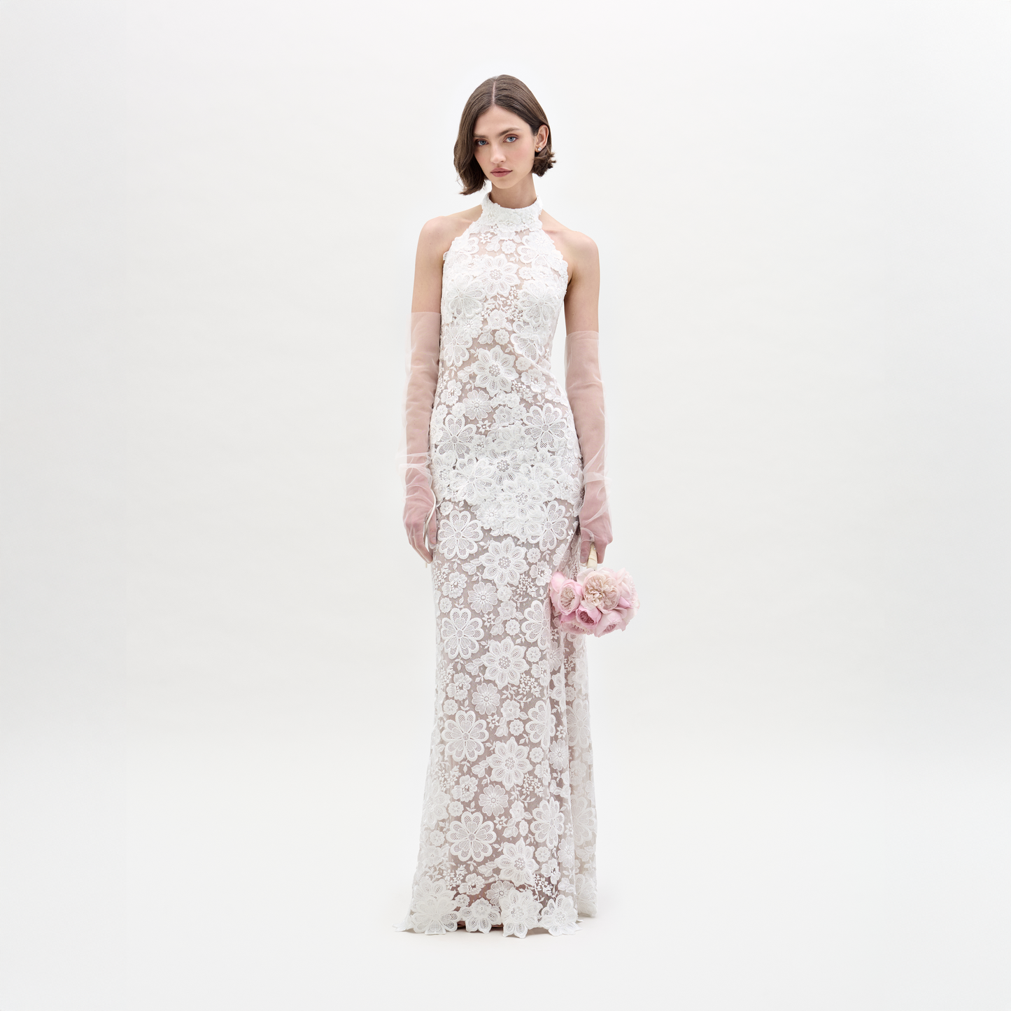 Una mujer con el Vestido Gardenia —un vestido largo blanco sin mangas con encaje transparente— sostiene flores rosas. Luce guantes largos y transparentes como complemento y se encuentra de pie frente a un fondo blanco liso, mirando a la cámara.