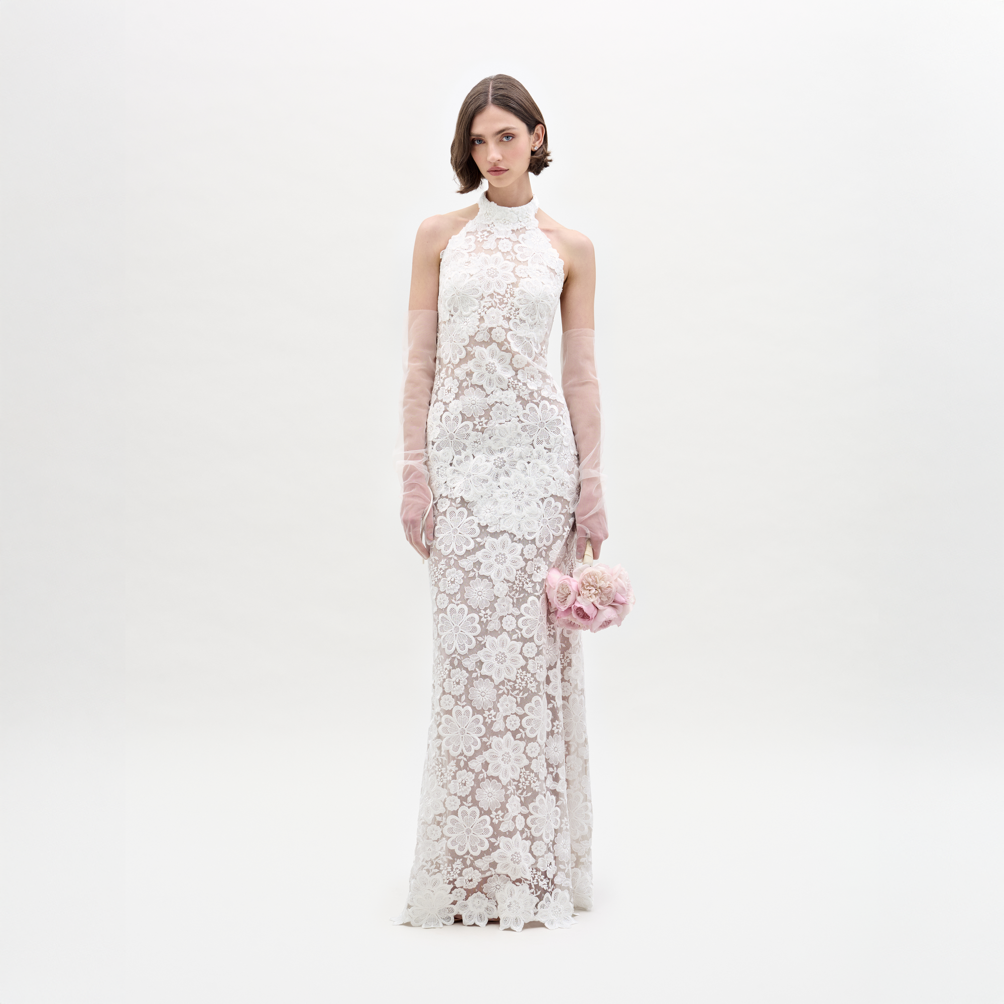 Una mujer con el Vestido Gardenia —un vestido largo blanco sin mangas con encaje transparente— sostiene flores rosas. Luce guantes largos y transparentes como complemento y se encuentra de pie frente a un fondo blanco liso, mirando a la cámara.