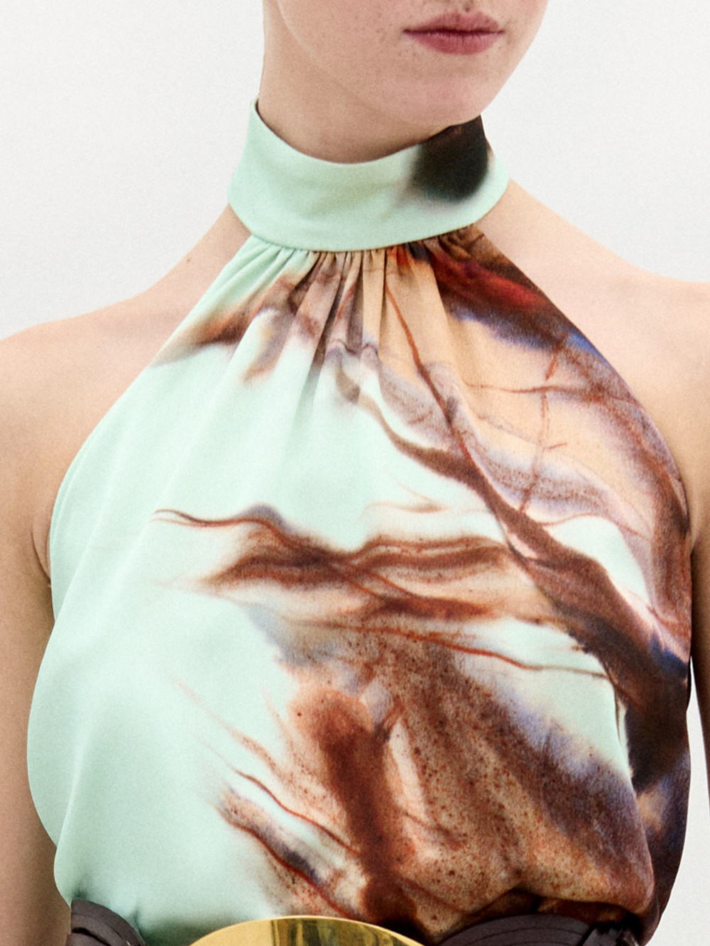 Una persona lleva la blusa Geovanna, una blusa sin mangas y de cuello alto, de color verde claro con motivos abstractos marrones, rojos y jaspeados oscuros. El rostro se muestra de la nariz hacia abajo. Confeccionada en Colombia con tela italiana.