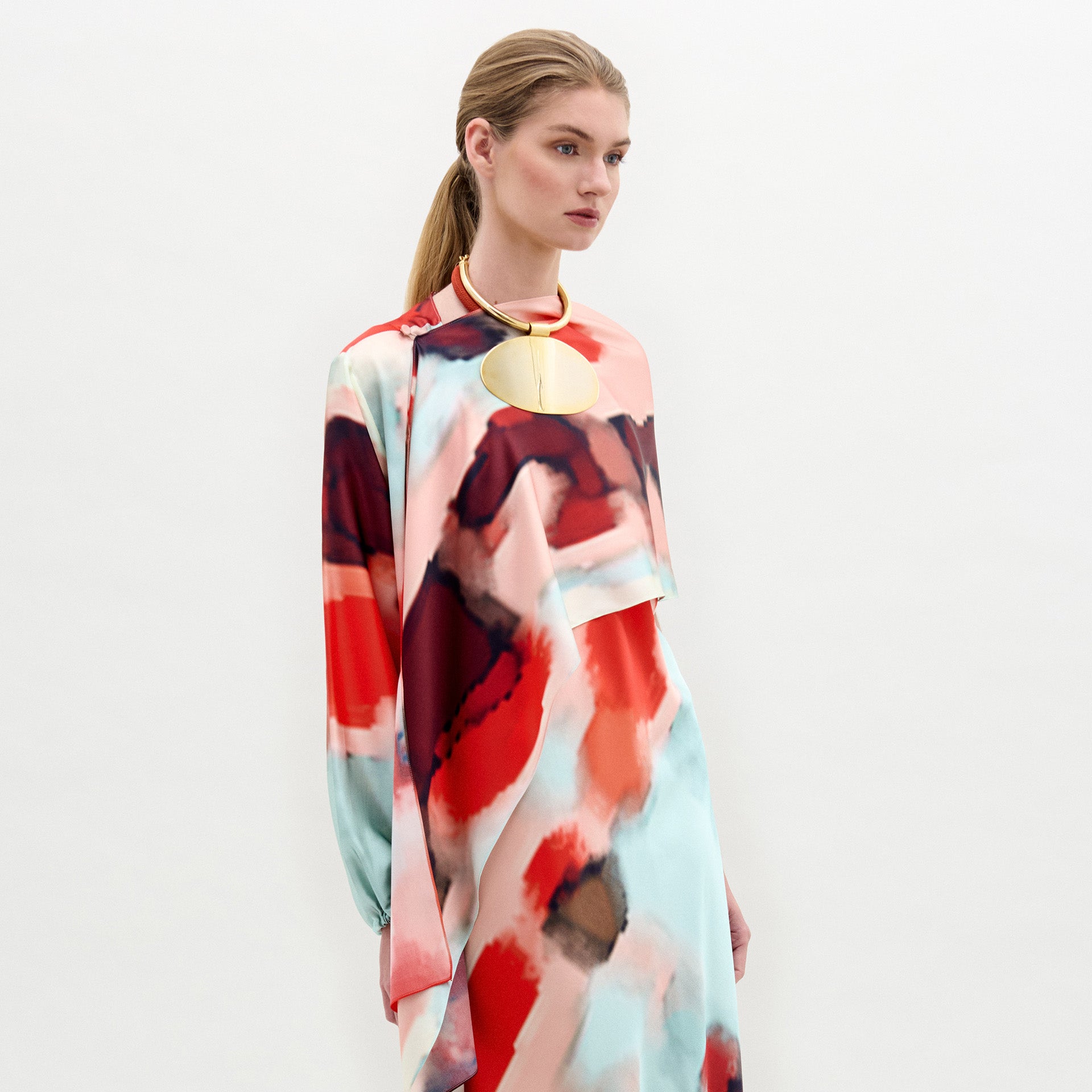 Una mujer con el pelo claro recogido luce el Gery Shrug, un vestido de manga larga con un motivo abstracto rojo, negro y azul claro en tela italiana, combinado con un gran collar circular dorado sobre un fondo blanco liso.