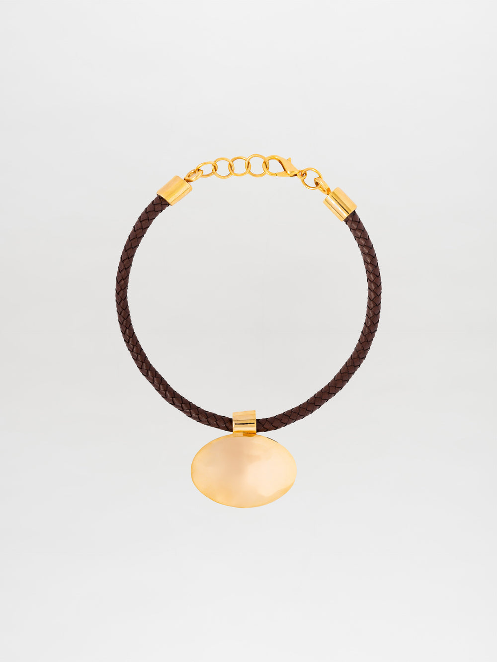 El Collar Gimena es una gargantilla de cuero trenzado marrón con cadena y broche de oro, elaborada en Colombia. Presenta un gran colgante ovalado de oro en el centro, sobre un fondo liso de color claro.