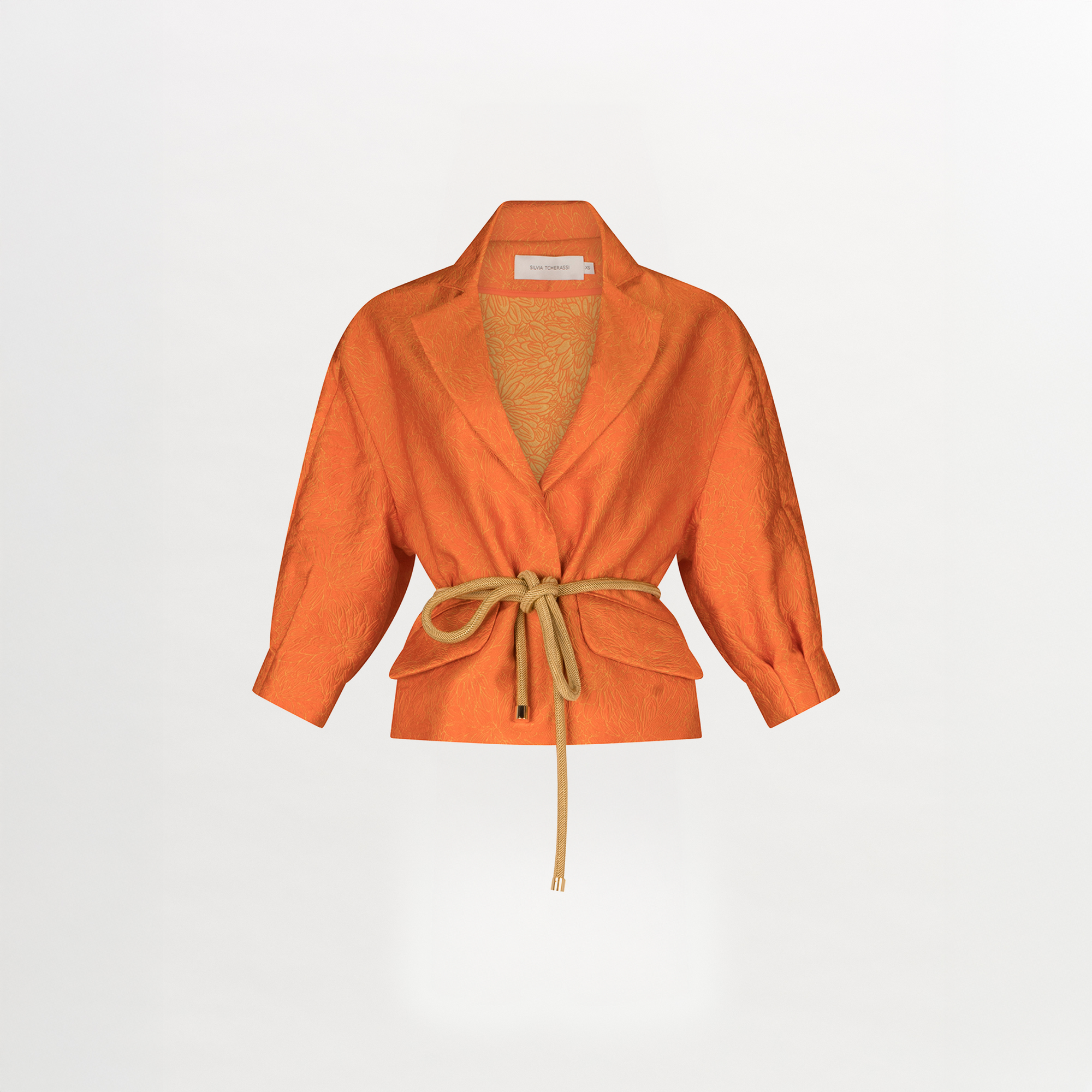 El Pantalón Moad se muestra sobre un fondo blanco; esta pieza corta de textura jacquard de color naranja presenta un cuello ancho, mangas tres cuartos y un cinturón de cuerda color canela atado a la cintura.