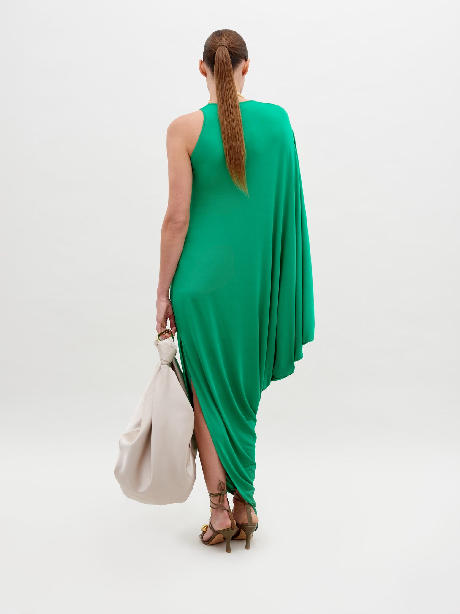 A person stands against a plain background, wearing a Golda Dress Green with gold accessories, holding a large beige handbag. Pre-order now for shipping by November 15th, 2024.