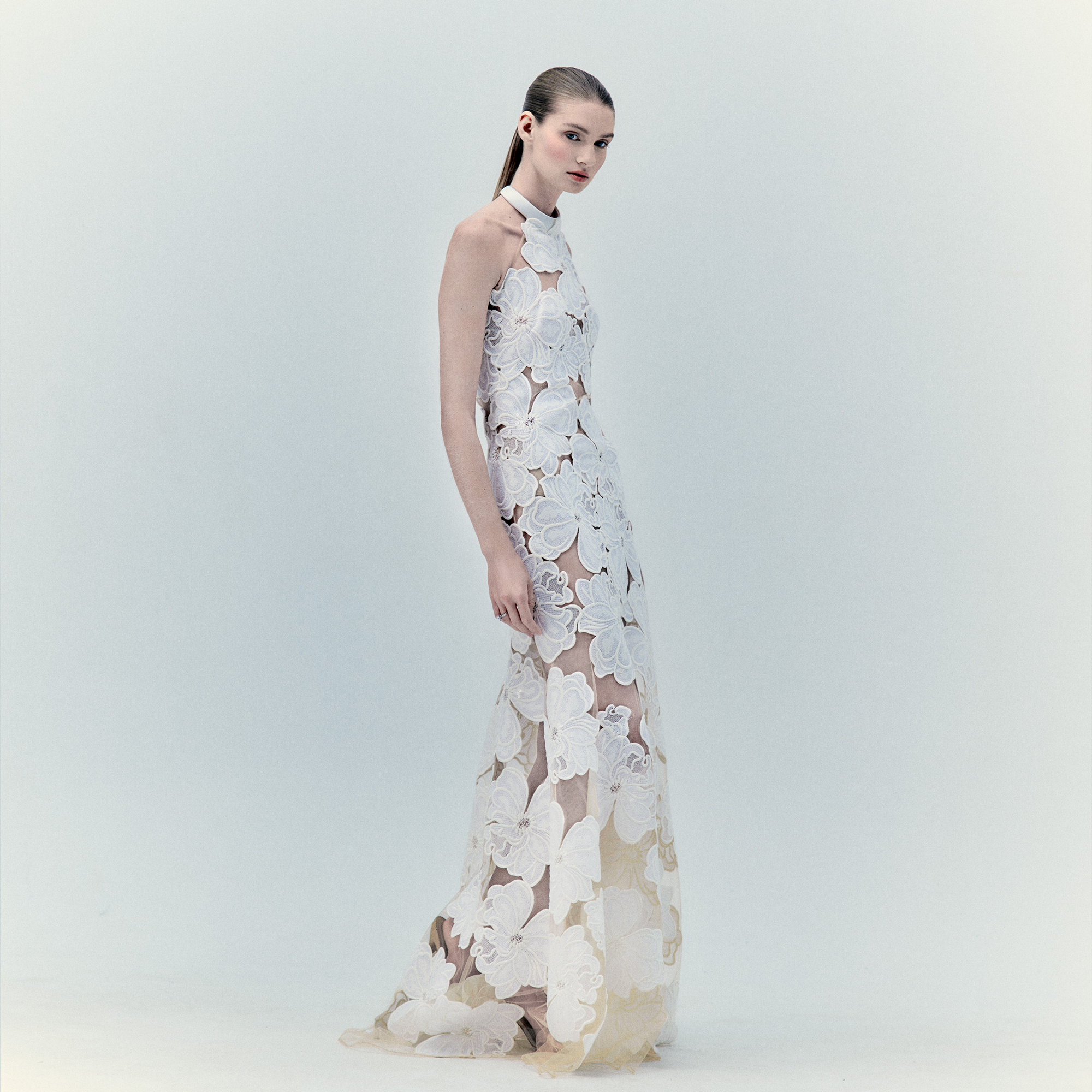 Una mujer aparece en un estudio gris claro con el Vestido Grace Pearl, un vestido largo y transparente con apliques florales y un elegante escote halter. Lleva el cabello liso peinado hacia atrás mientras mira a la cámara con una expresión neutra.