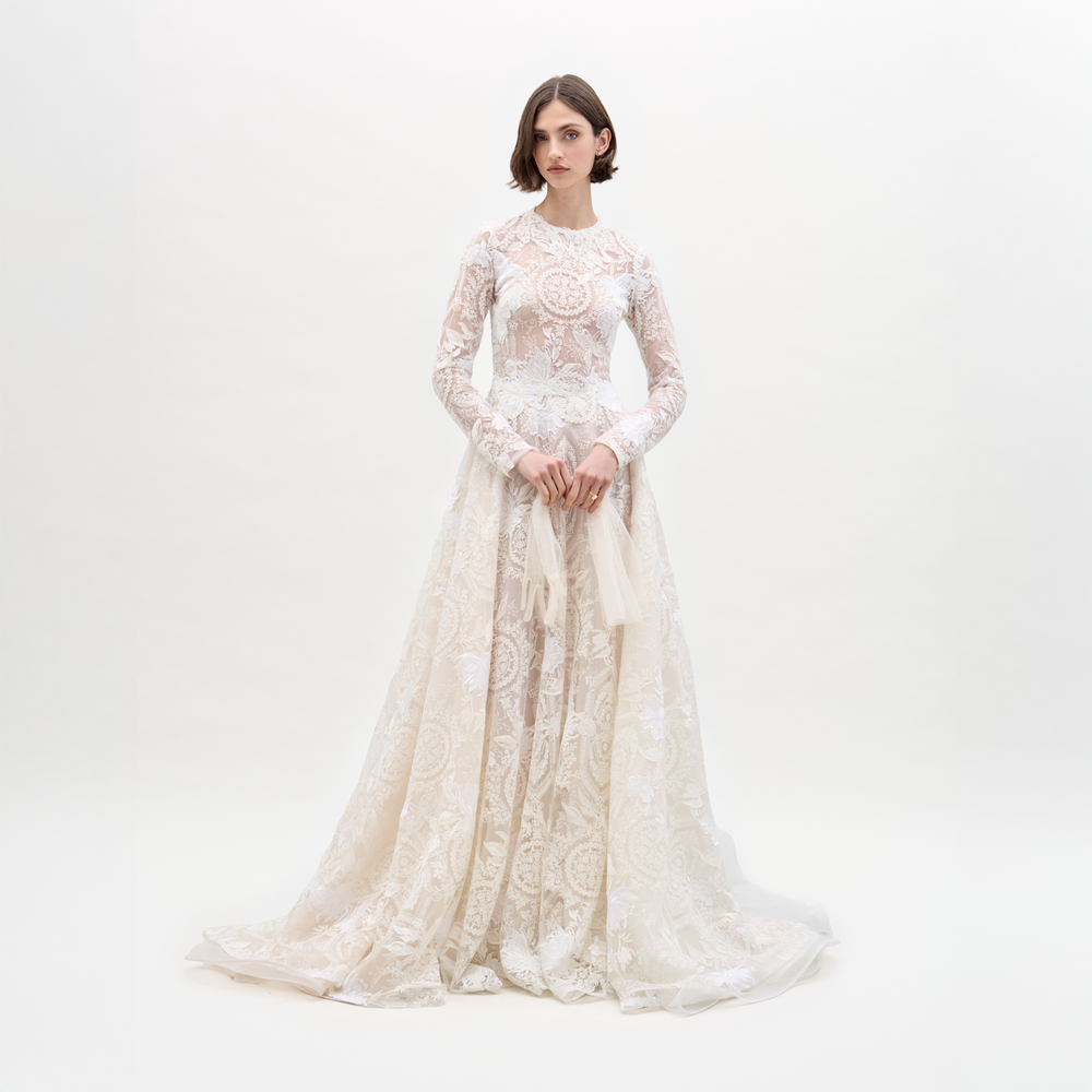 Una mujer mira hacia adelante con un vestido Hermione largo y de manga larga, con encaje translúcido y corpiño ajustado, sosteniendo un velo transparente. El vestido blanco de encaje tiene una falda fluida y se presenta sobre un fondo blanco liso.