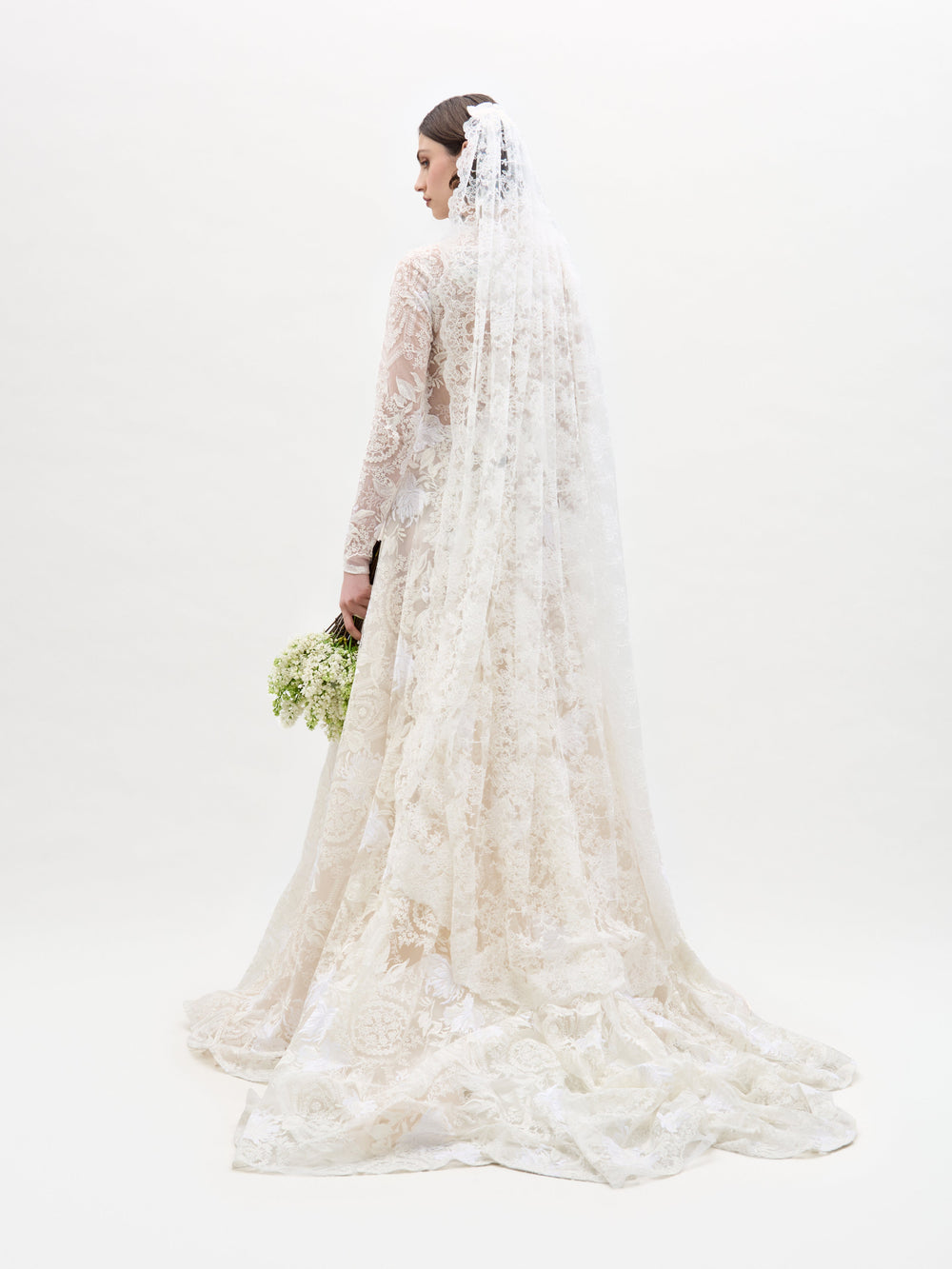 Una novia, de espaldas, luce el vestido Hermione: un elegante vestido de novia largo de encaje con velo a juego. Sostiene un ramo de flores blancas y verdes sobre un fondo blanco liso.