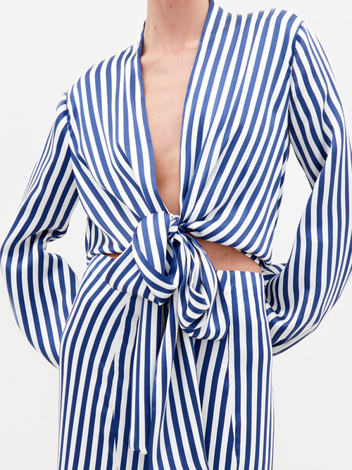 A person stands in the Honey Blouse White Blue Stripes, featuring a tie at the waist and a ruffled hem, paired with embellished sandals. This stylish ensemble is available for pre-order with shipping by February 15th.