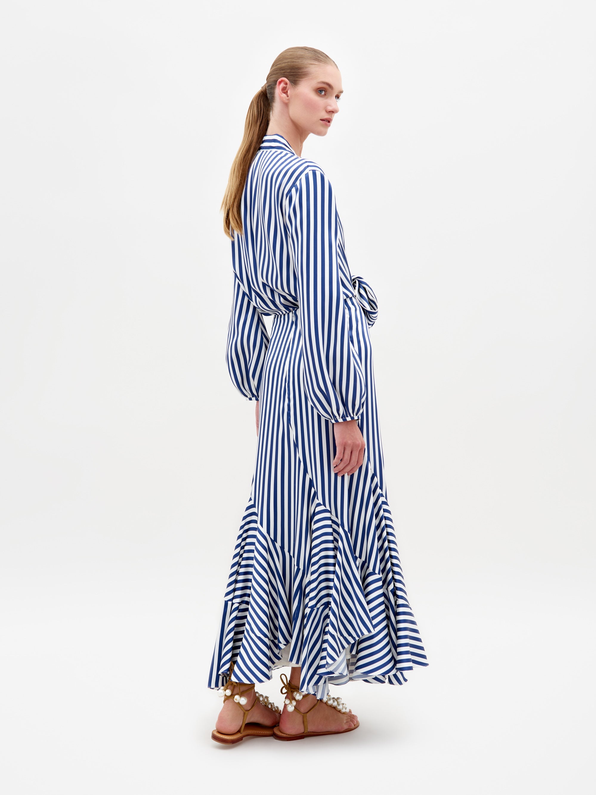 Una persona con cabello largo luce un elegante vestido de rayas azules y blancas de Falda Florina confeccionado en un lujoso textil italiano, con cintura ceñida y falda con volados. Junto con sandalias marrones, está parada de lado sobre un fondo blanco liso, mirando hacia atrás con gracia.