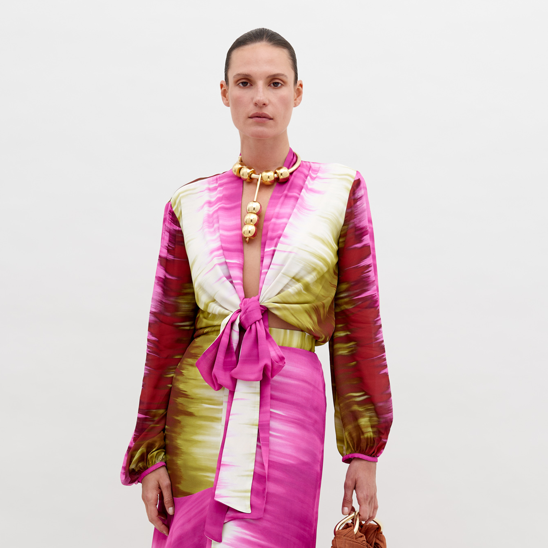 Una mujer, sobre un fondo blanco liso, lleva la Blusa Honey, una blusa de manga larga con estampado rosa y verde y lazo delantero -elaborada en textil italiano-, que complementa con un collar de cuentas doradas y un pequeño bolso marrón con textura.