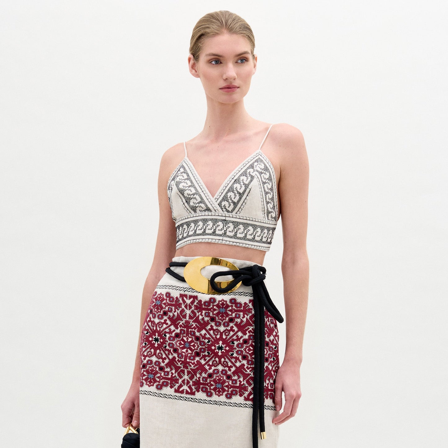 Una mujer con el pelo claro recogido en un moño bajo lleva el top Ibbie, un corpiño geométrico sin mangas en blanco y negro combinado con una falda roja estampada. Se complementa con un cinturón ancho negro y dorado y un bolso pequeño.