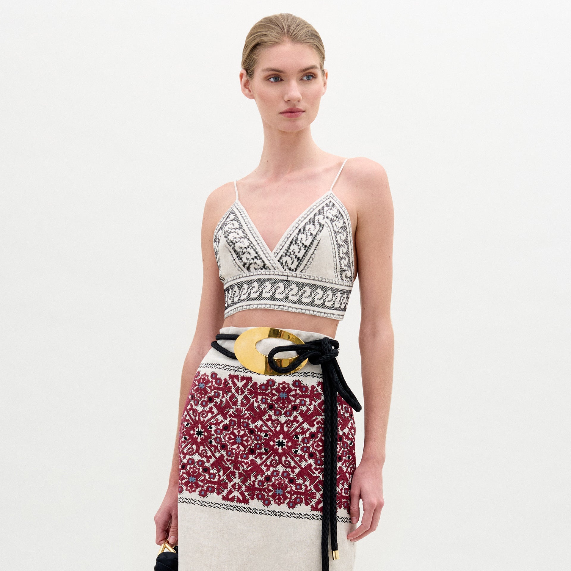 Una mujer con el pelo claro recogido en un moño bajo lleva el top Ibbie, un corpiño geométrico sin mangas en blanco y negro combinado con una falda roja estampada. Se complementa con un cinturón ancho negro y dorado y un bolso pequeño.