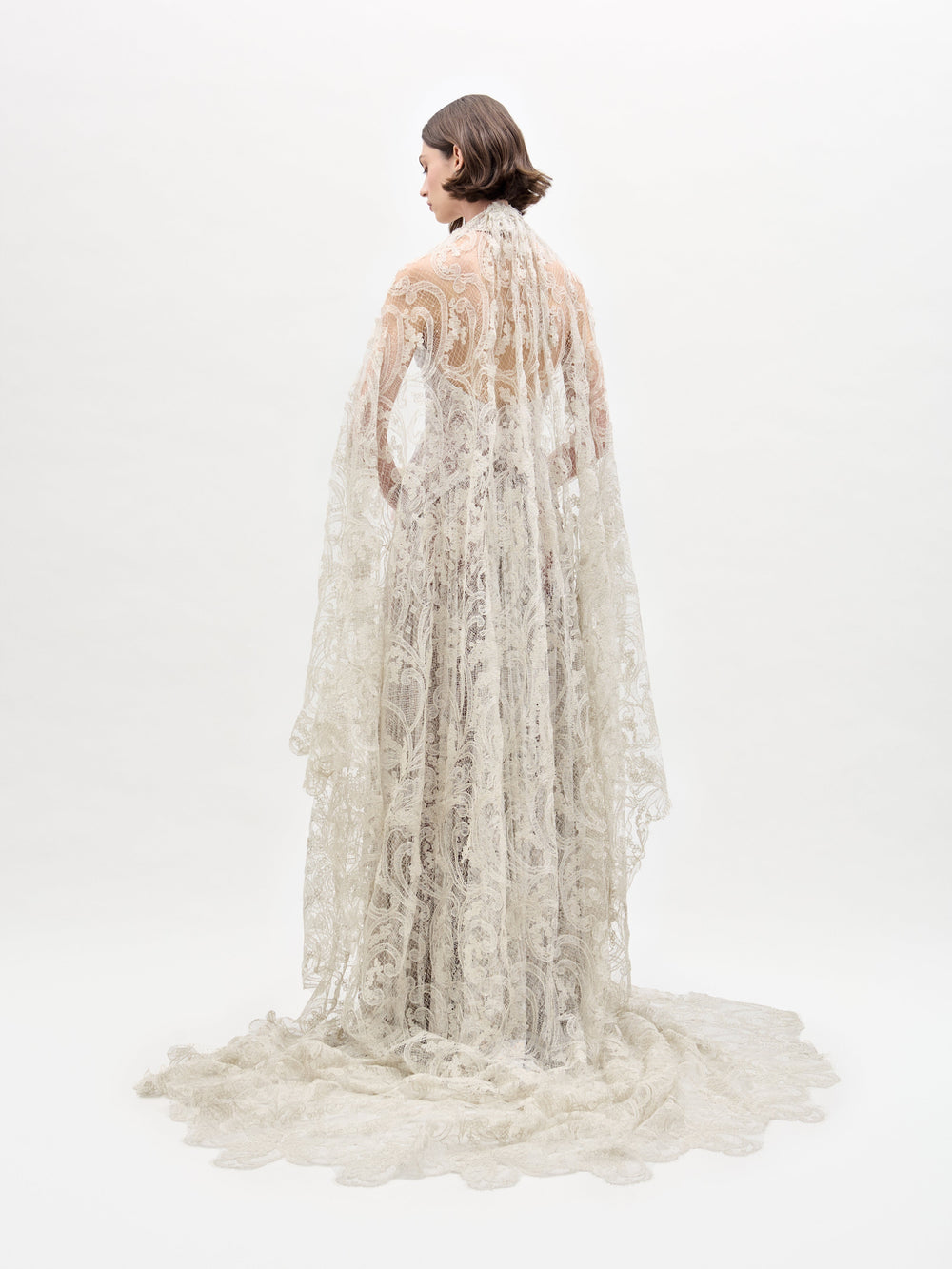 El vestido Ilana luce un intrincado diseño de encaje hasta el suelo, con una larga cola y elaborados patrones. Una persona aparece de espaldas a él sobre un fondo blanco liso; su cabello corto y oscuro completa el elegante look.