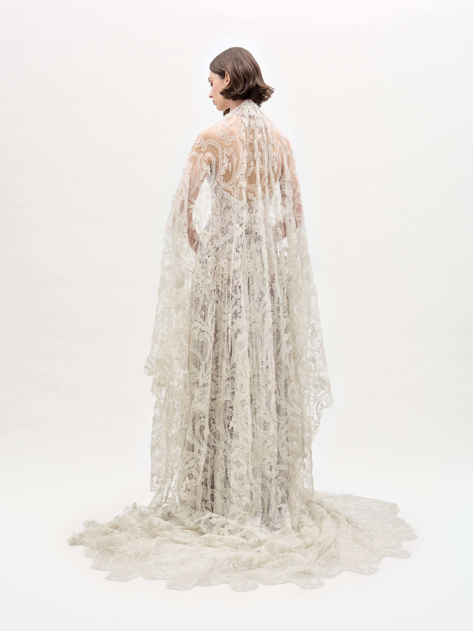 El vestido Ilana luce un intrincado diseño de encaje hasta el suelo, con una larga cola y elaborados patrones. Una persona aparece de espaldas a él sobre un fondo blanco liso; su cabello corto y oscuro completa el elegante look.