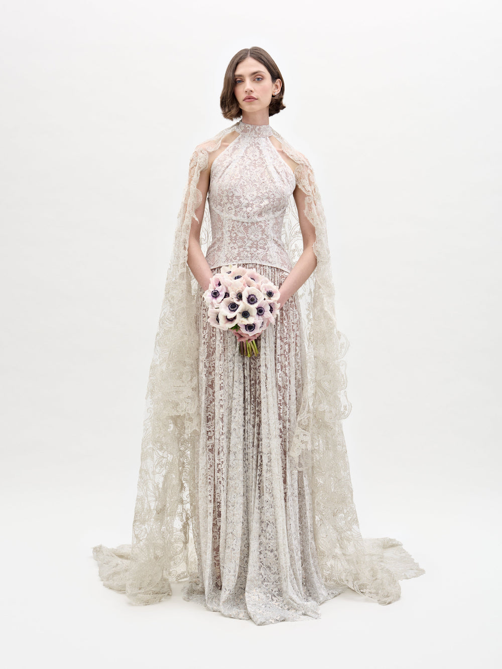 Una mujer con el vestido Ilana, un vestido de encaje con intrincados estampados, cuello alto y capa transparente, se encuentra de pie sobre un fondo blanco liso sosteniendo un ramo de flores claras. Su cabello es corto y de peinado sencillo.