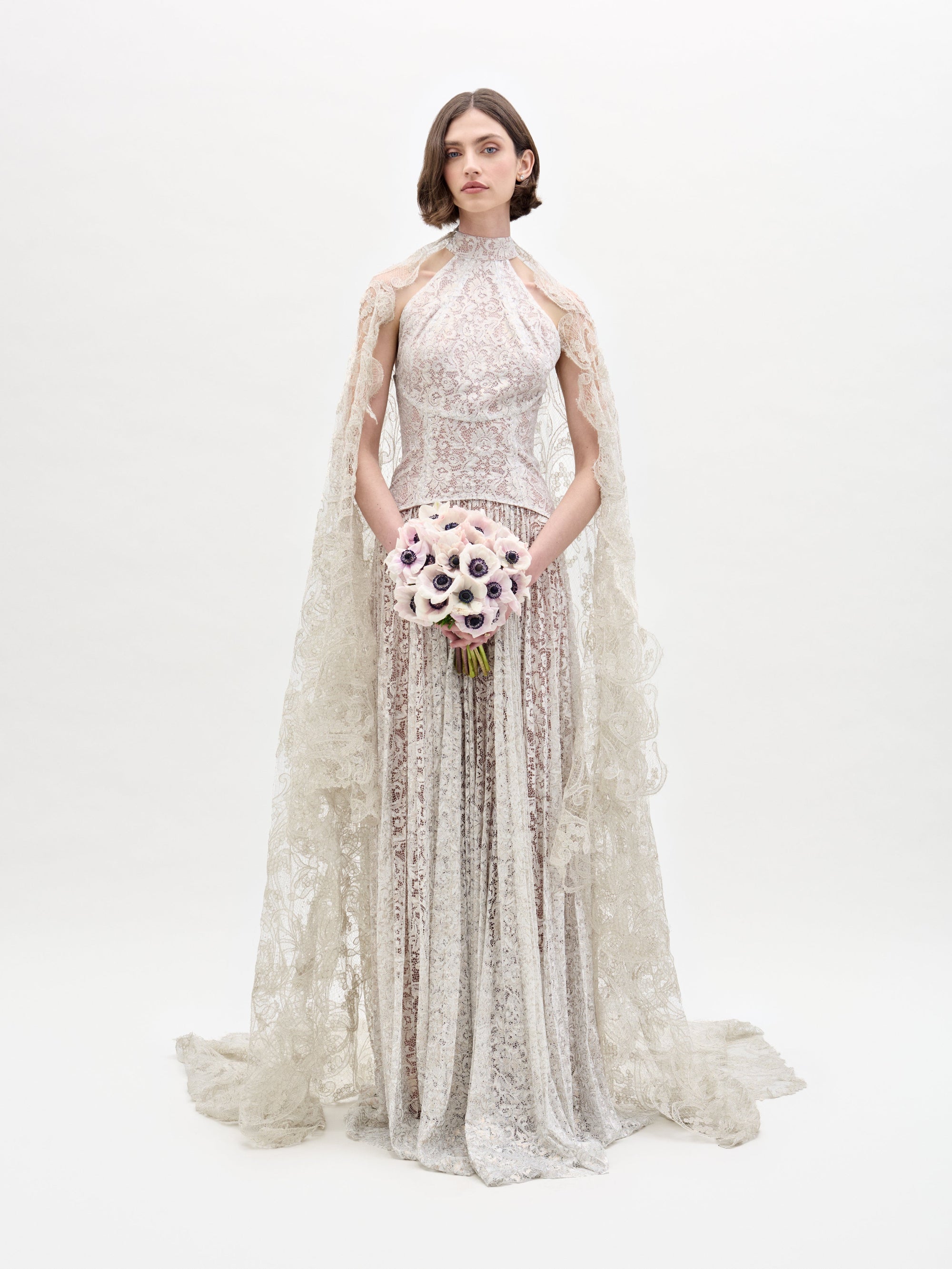 Una mujer con el vestido Ilana, un vestido de encaje con intrincados estampados, cuello alto y capa transparente, se encuentra de pie sobre un fondo blanco liso sosteniendo un ramo de flores claras. Su cabello es corto y de peinado sencillo.