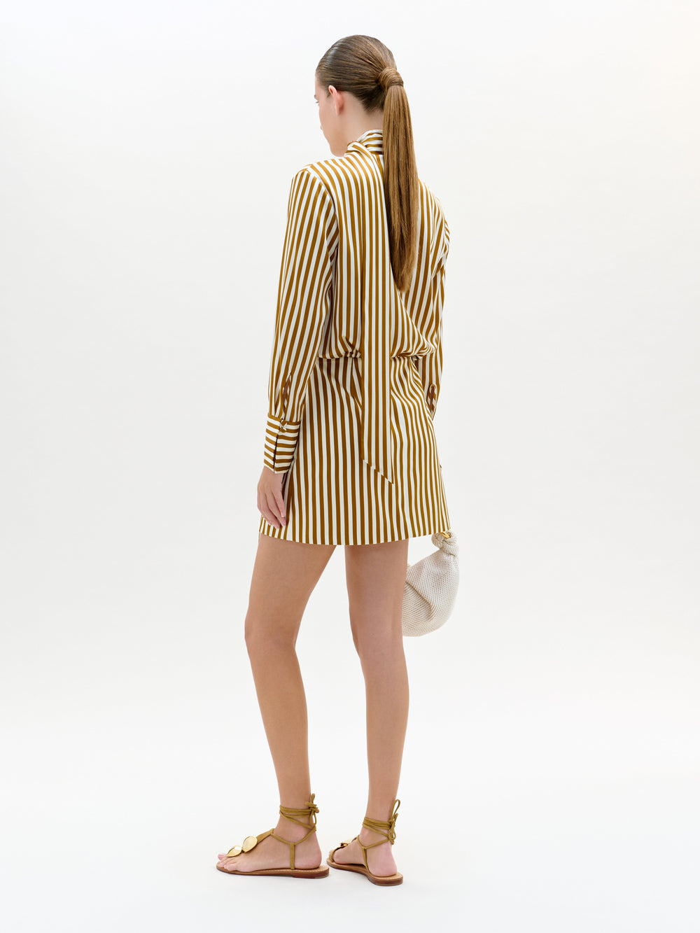 Una mujer mira hacia otro lado y lleva el vestido Imelda, un minivestido de manga larga a rayas verticales amarillas y marrones con detalle de pañuelo. Lleva un pequeño bolso blanco, calza sandalias planas marrones y está de pie sobre un fondo claro y liso.