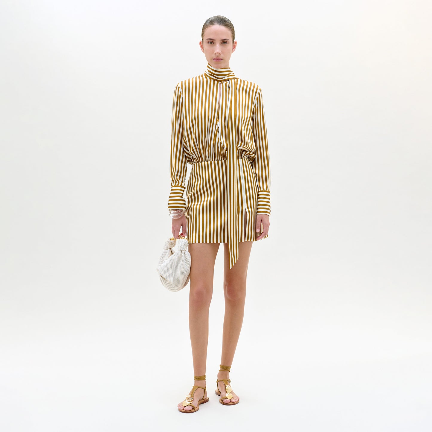 Sobre un fondo blanco, una persona lleva el vestido Imelda, un minivestido amarillo y blanco a rayas verticales con cuello alto y detalle de lazo, combinado con sandalias doradas y un pequeño bolso blanco.