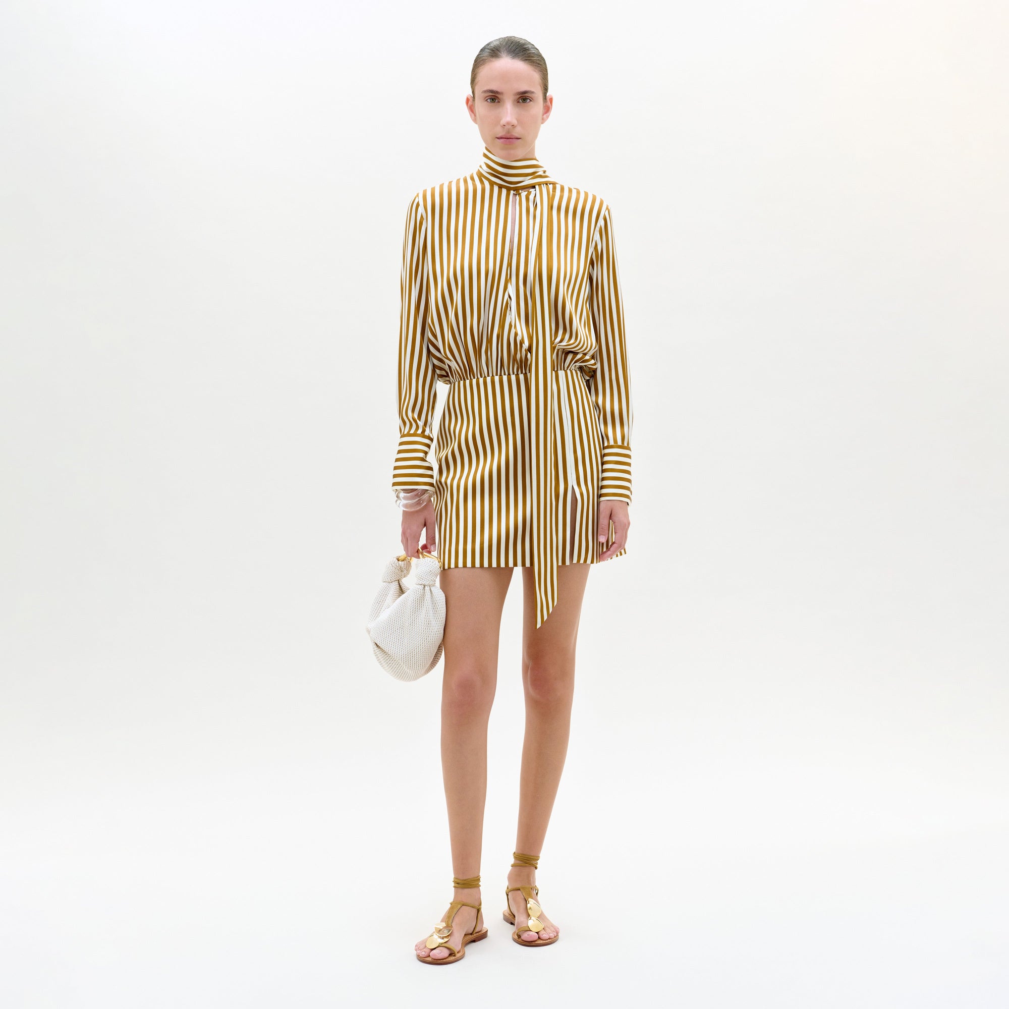 Sobre un fondo blanco, una persona lleva el vestido Imelda, un minivestido amarillo y blanco a rayas verticales con cuello alto y detalle de lazo, combinado con sandalias doradas y un pequeño bolso blanco.